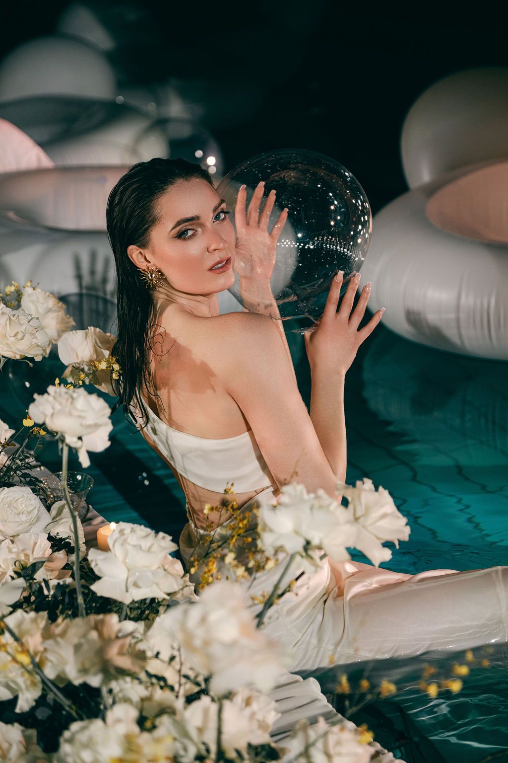 Woman in white swimsuit holding a disco ball in a pool surrounded by white flowers and inflatable rings.