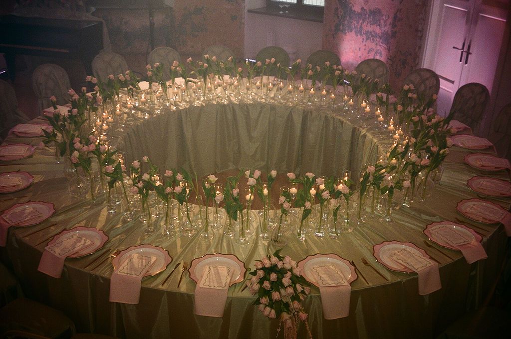 Elegant round wedding reception table with candles, pink roses, and gold place settings in a softly lit venue.