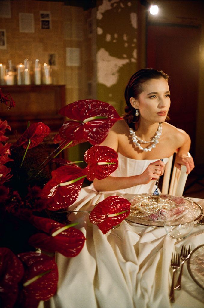Elegant bride in white strapless gown seated at a table with red anthurium flowers and pearl necklace.