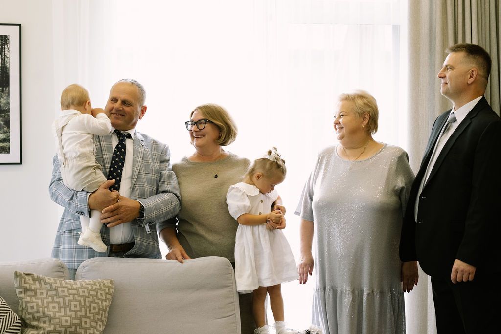 Family gathering with grandparents holding babies and toddler in elegant attire indoors.