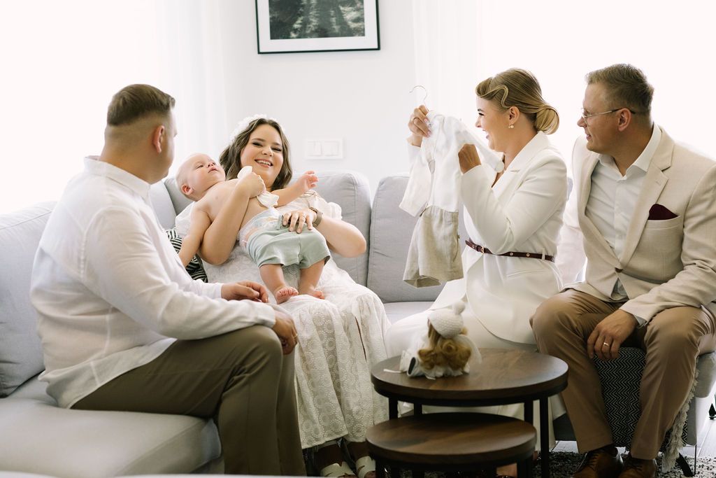 Family gathering on a sofa, admiring a newborn baby in a bright, cozy living room setting.