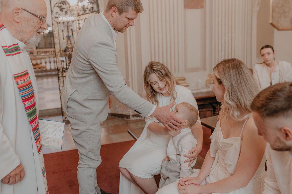 A family gathered in a church for a baptism ceremony, with a priest blessing a baby held by its mother.