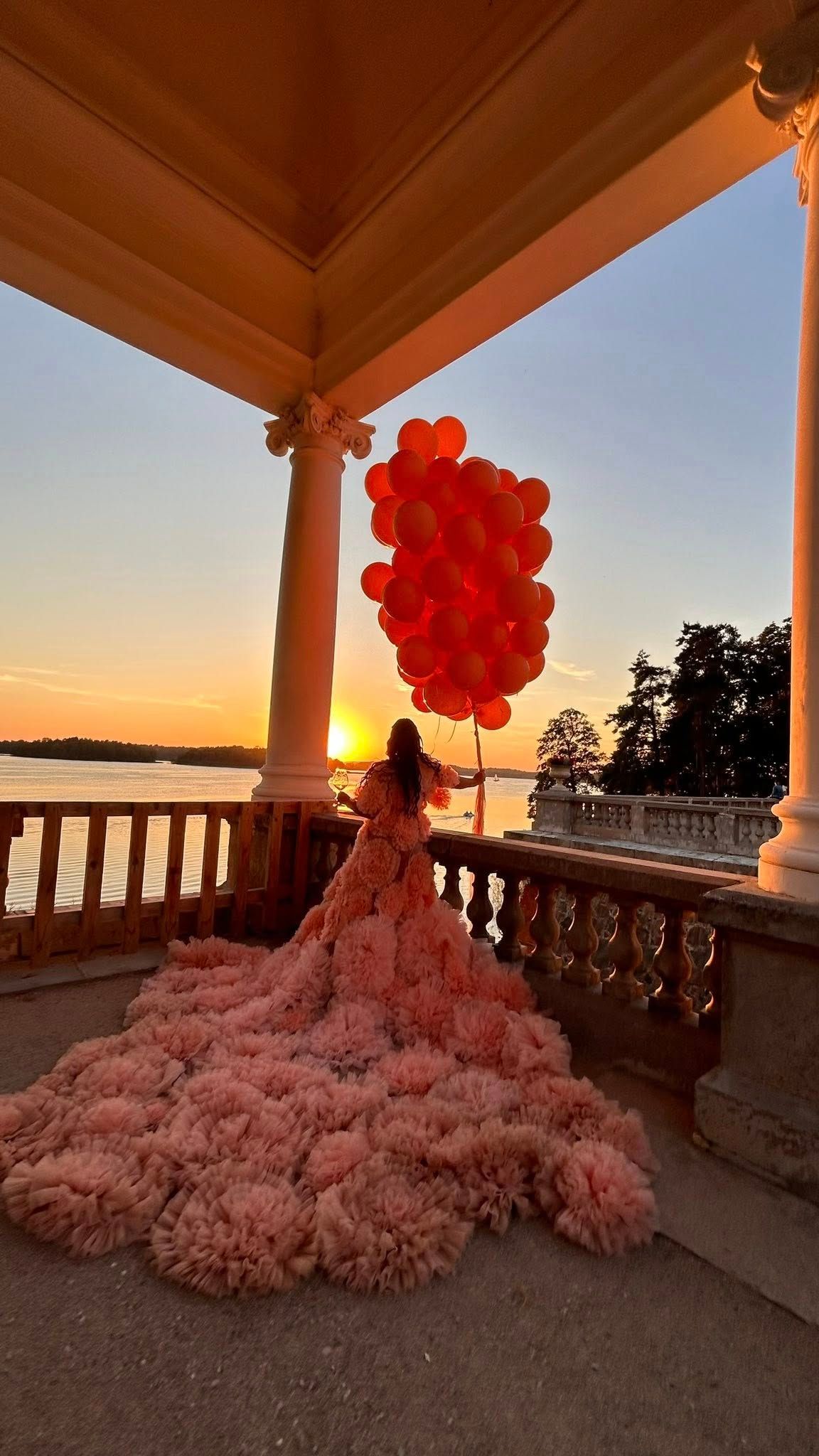 Woman in pink ruffled gown holds orange balloons on elegant balcony at sunset overlooking serene waterfront.