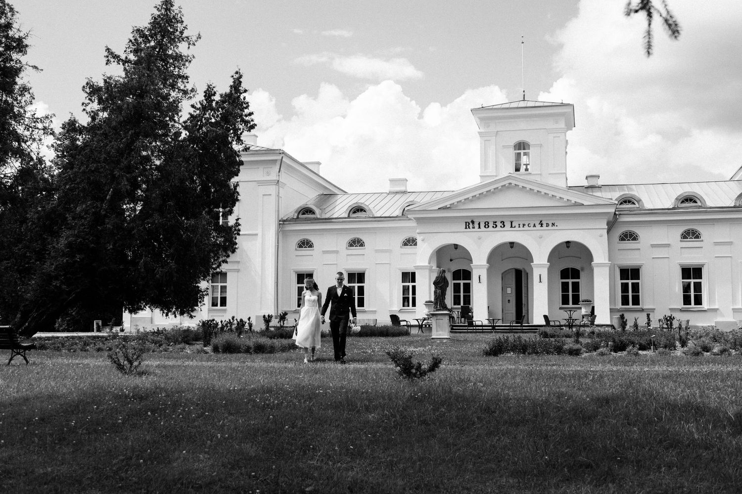 Black and white wedding photo of couple holding hands outside elegant white manor house with columns and lush gardens.