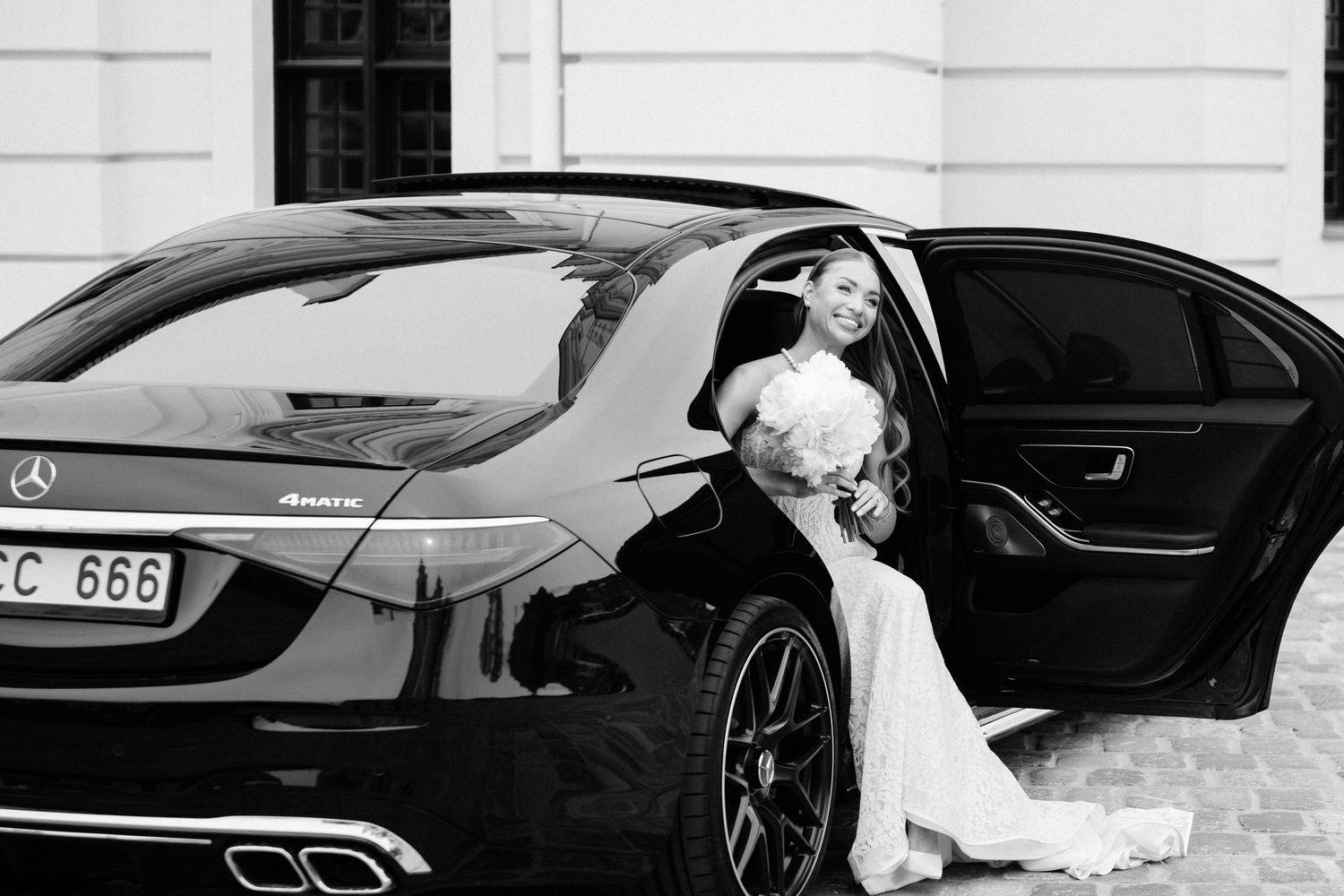 Bride in white wedding dress stepping out of a black Mercedes-Benz luxury car, holding a bouquet, black and white photo.
