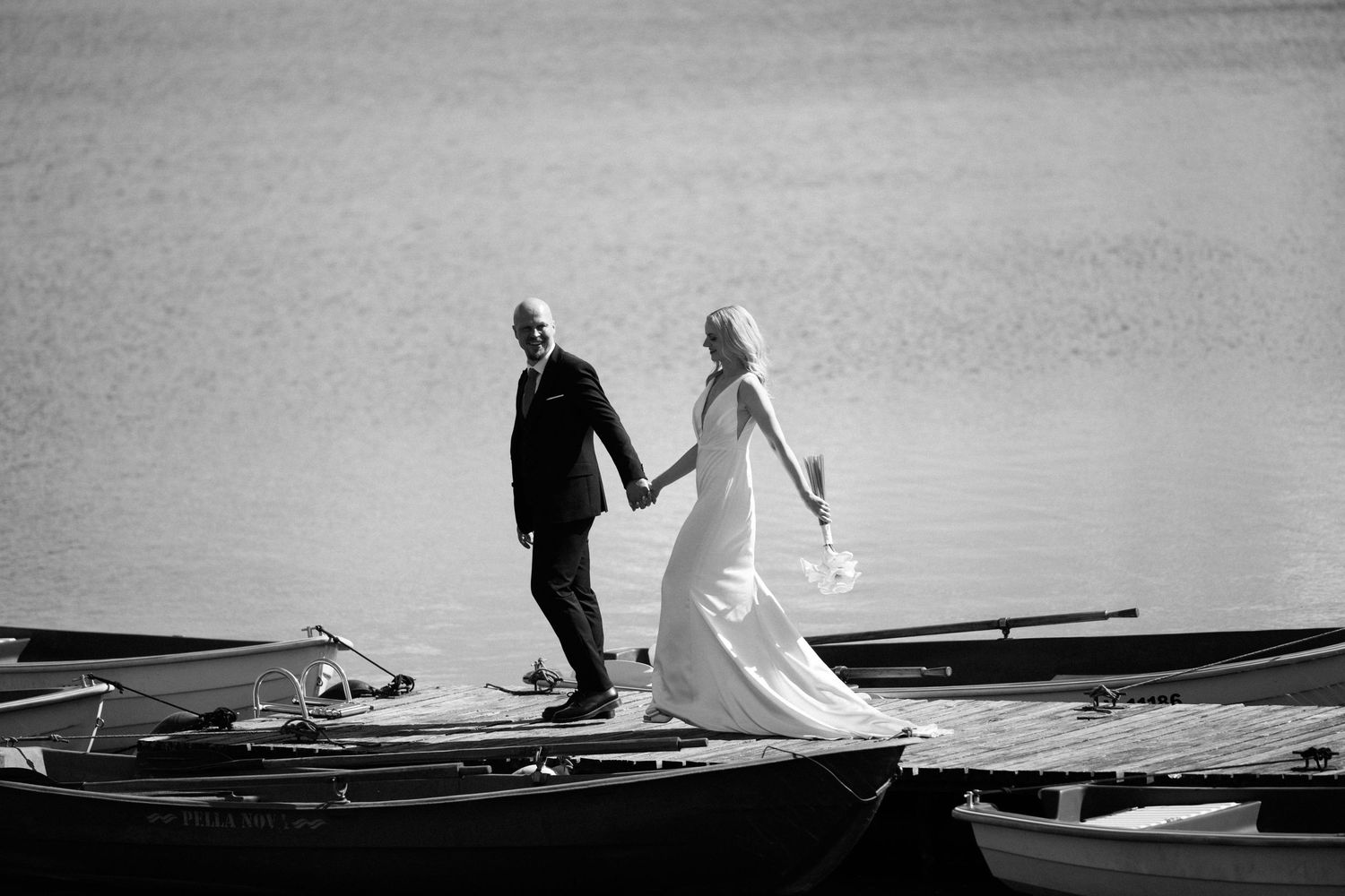 Bride in a flowing white gown and groom in a dark suit hold hands while walking along a wooden dock beside rowboats in black and white.