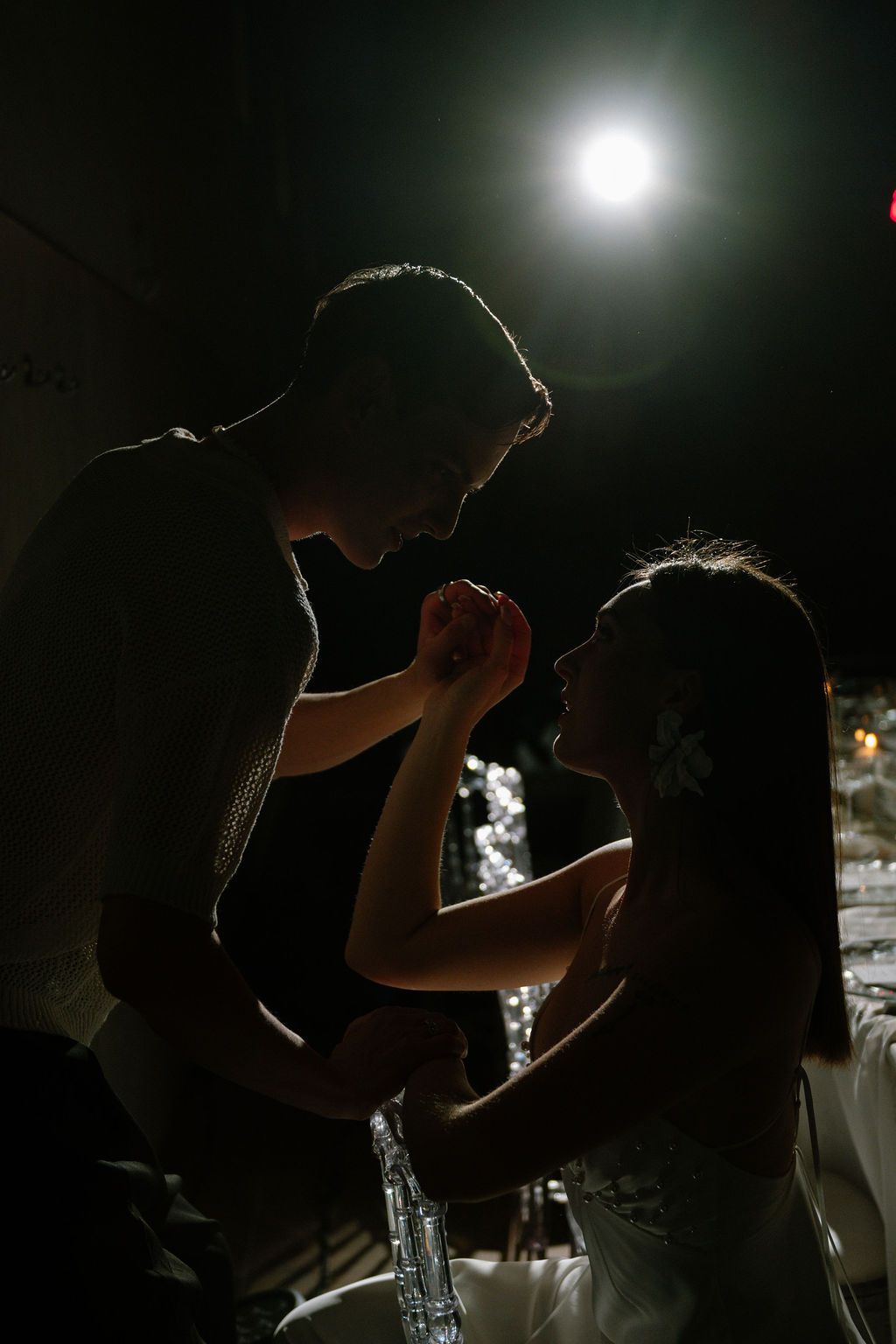 Silhouetted couple holding hands in a romantic moment, a bright spotlight glowing behind them in a dark reception setting.