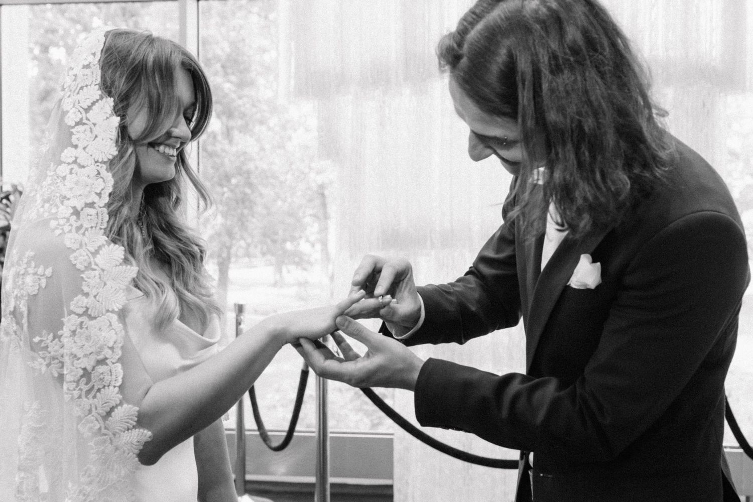 Black and white photo of a groom in a dark suit placing a wedding ring on a smiling bride's finger during the ceremony.