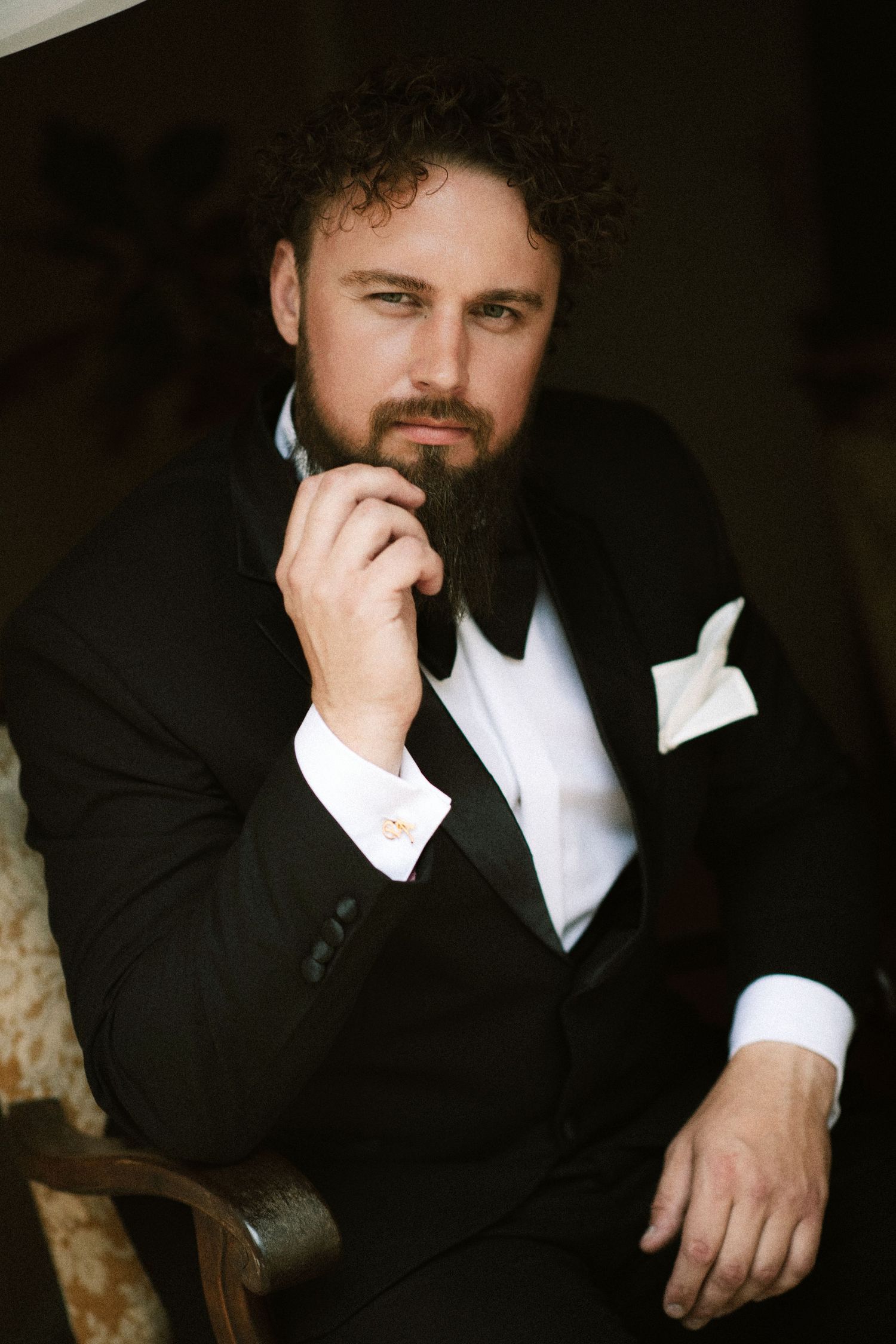 Elegant bearded groom in black tuxedo with white pocket square sitting thoughtfully in a chair.