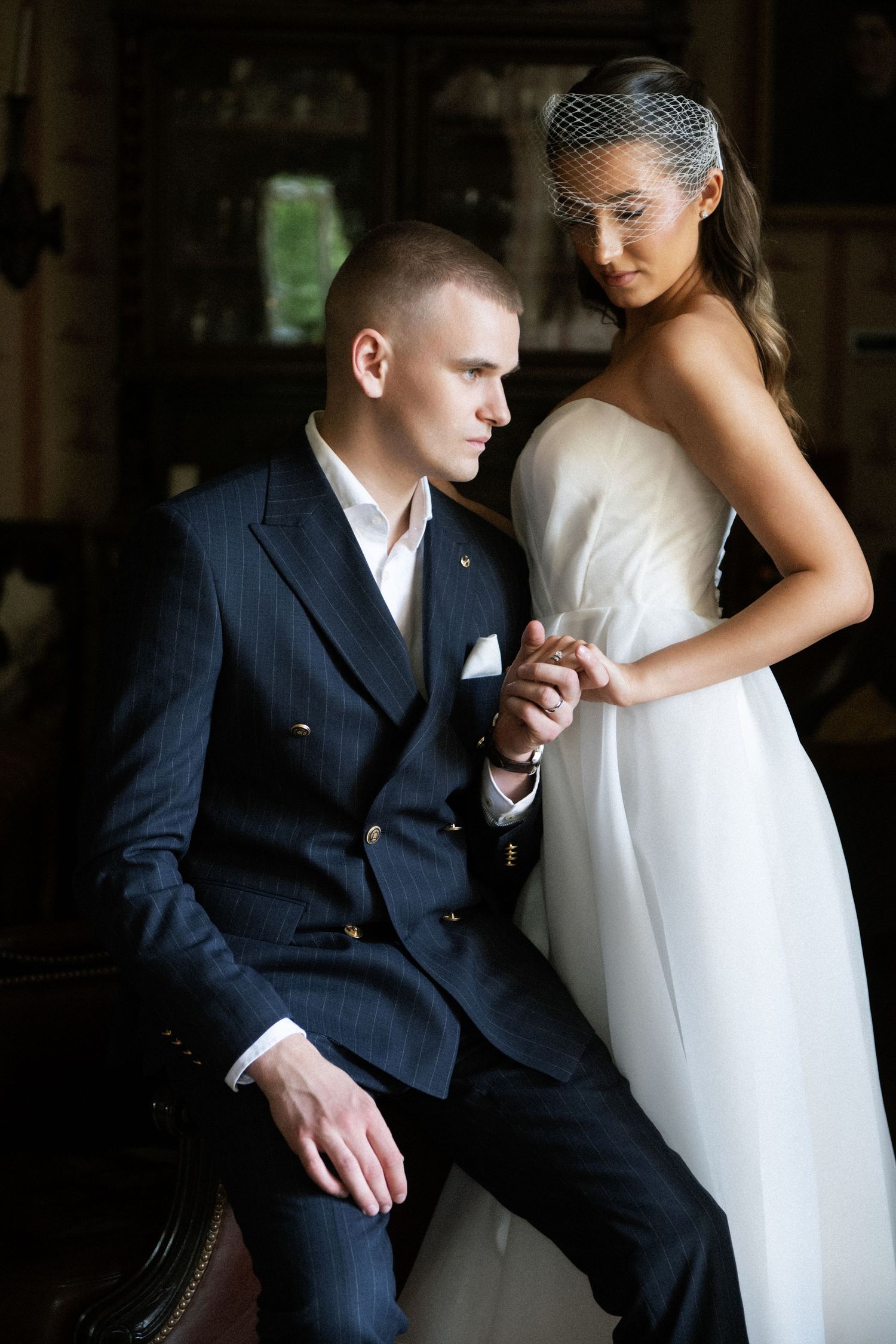 Elegant wedding couple posing indoors, groom in navy suit seated holding bride's hand in white gown and veil.