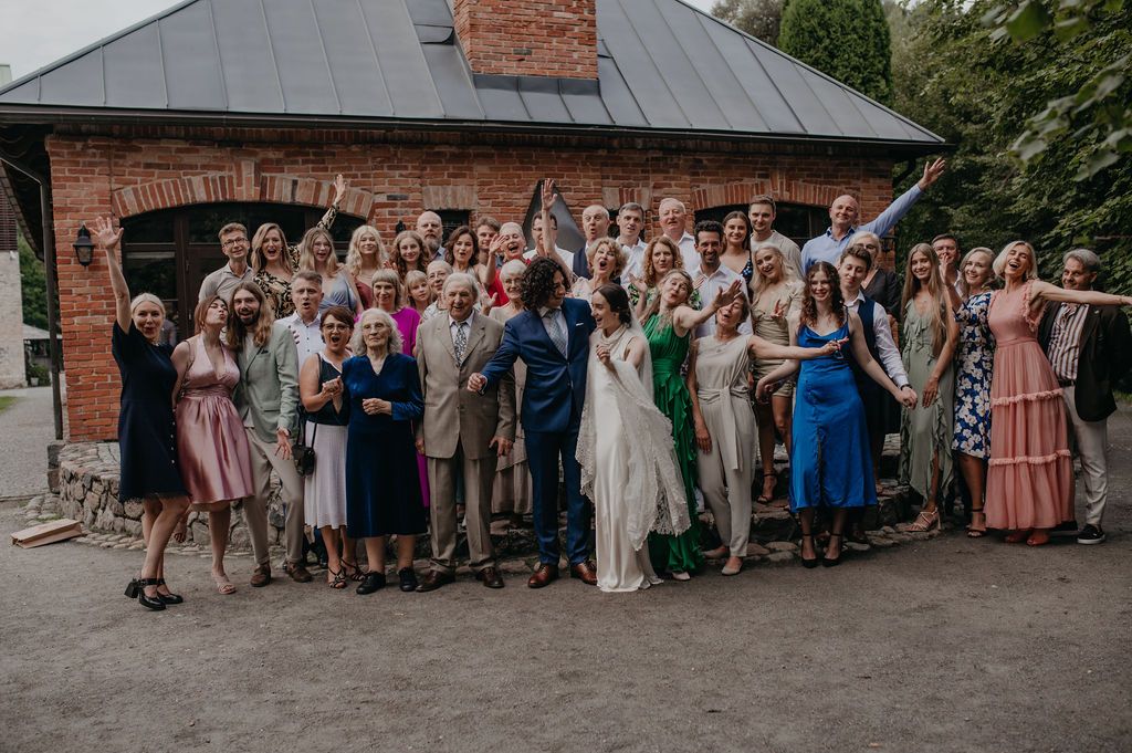 Large wedding group photo with bride and groom surrounded by guests outside a brick building.