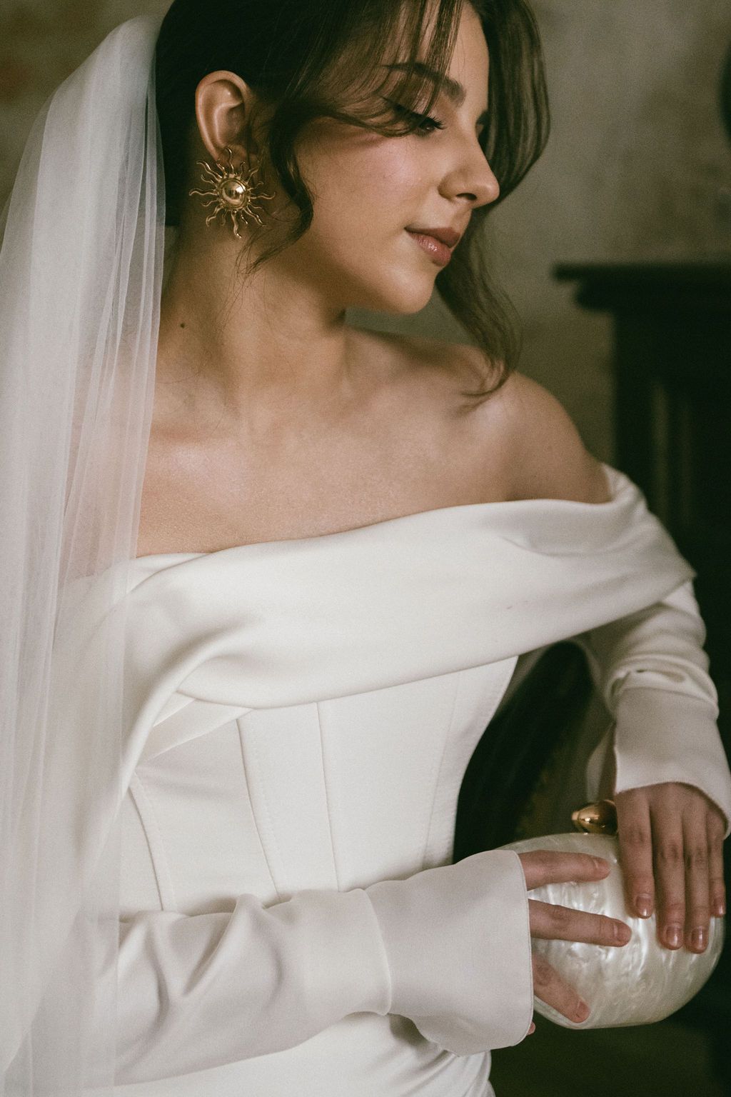 Bride in off-shoulder white satin gown with gold sunburst earrings and veil in elegant wedding portrait.