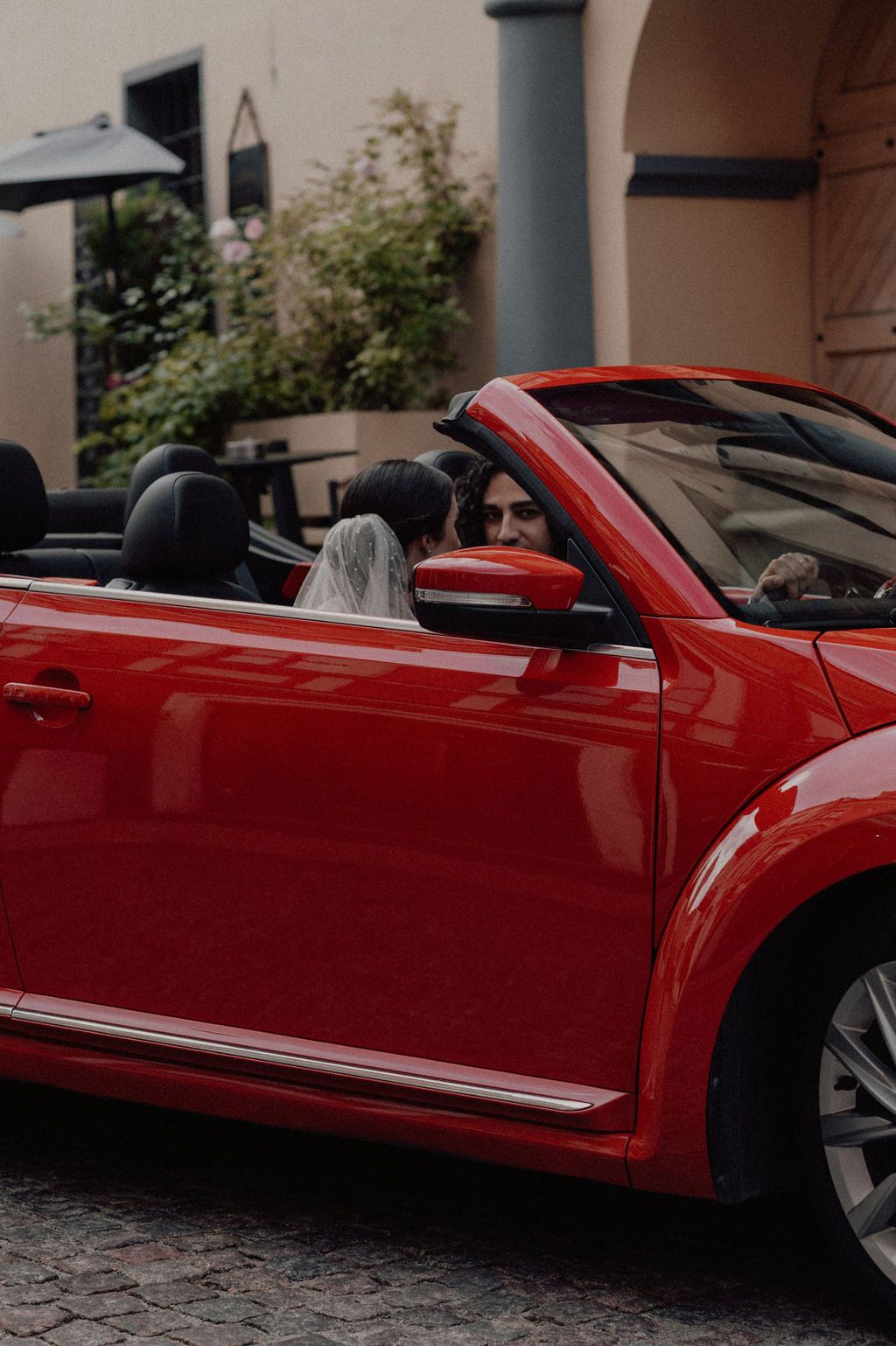 Newlywed couple sitting in a red convertible Volkswagen Beetle on their wedding day, sharing a romantic moment.