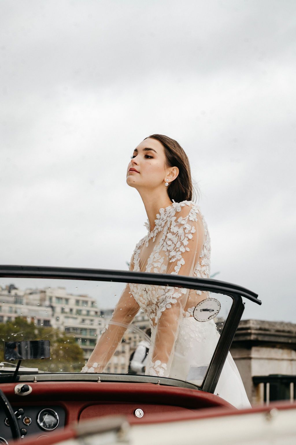 Bride in sheer lace gown standing in vintage convertible car in Paris.