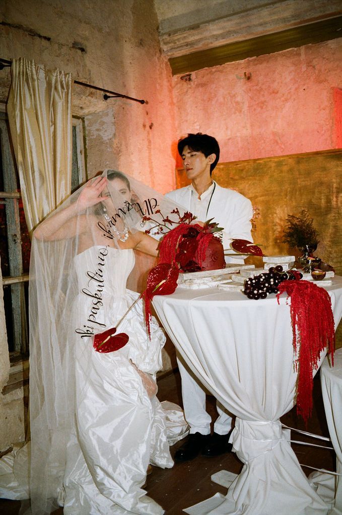 Couple in white wedding attire at a draped table with red floral decor in a rustic venue.