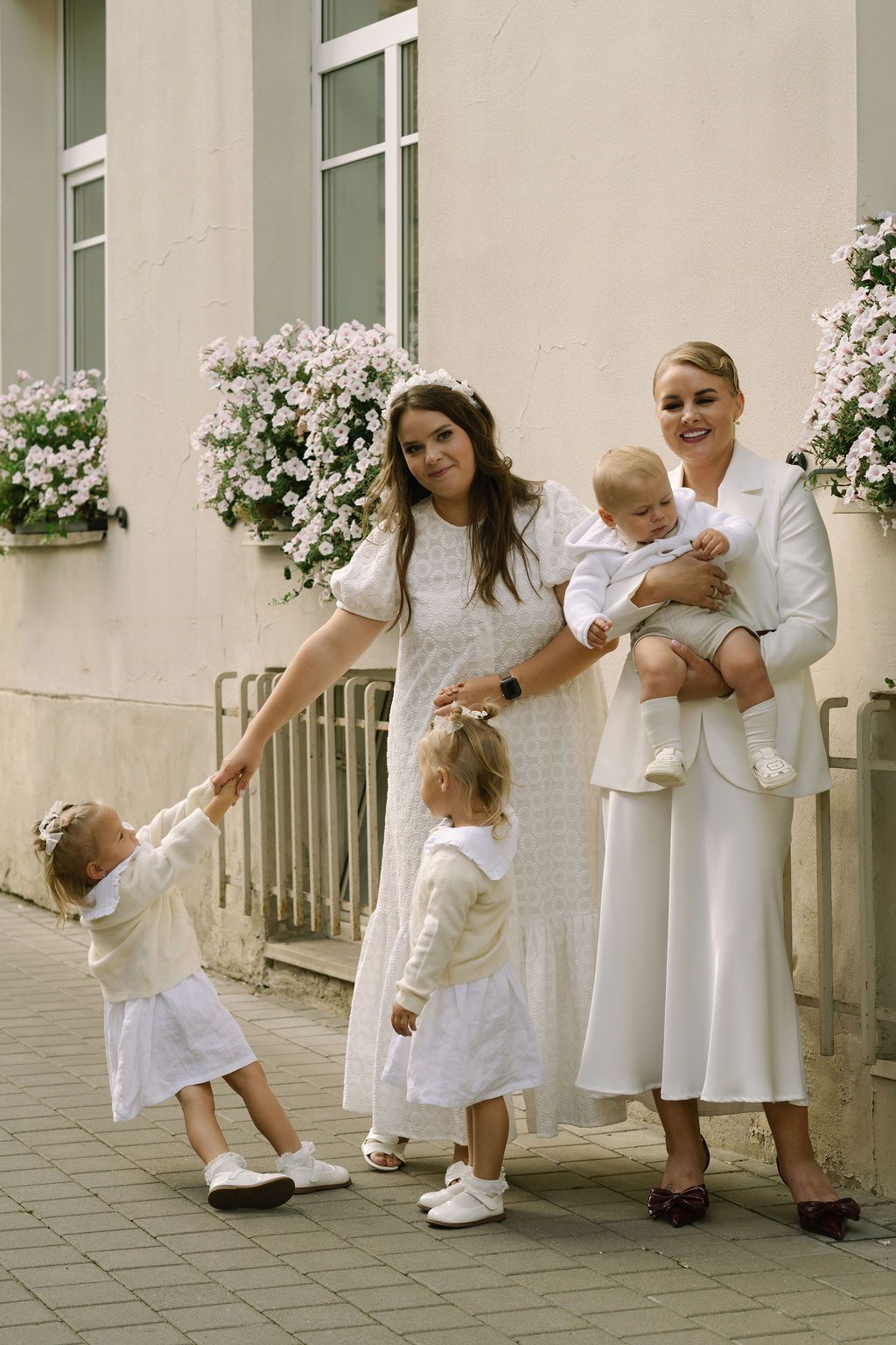 Two women in white outfits pose outdoors with three young children near a building with flower boxes.