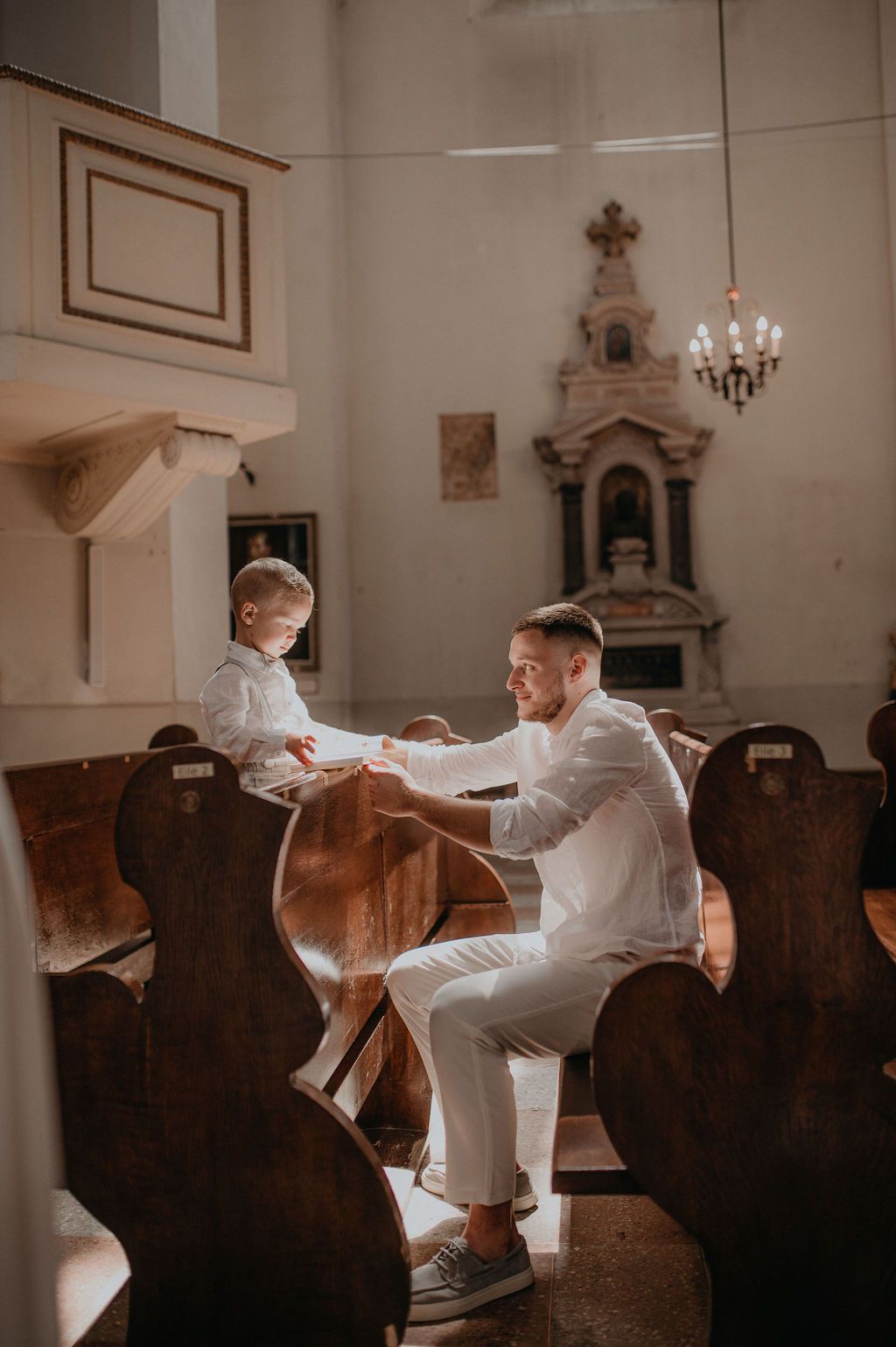 A smiling man in white attire interacts with a young boy in a sunlit historic church with wooden pews.