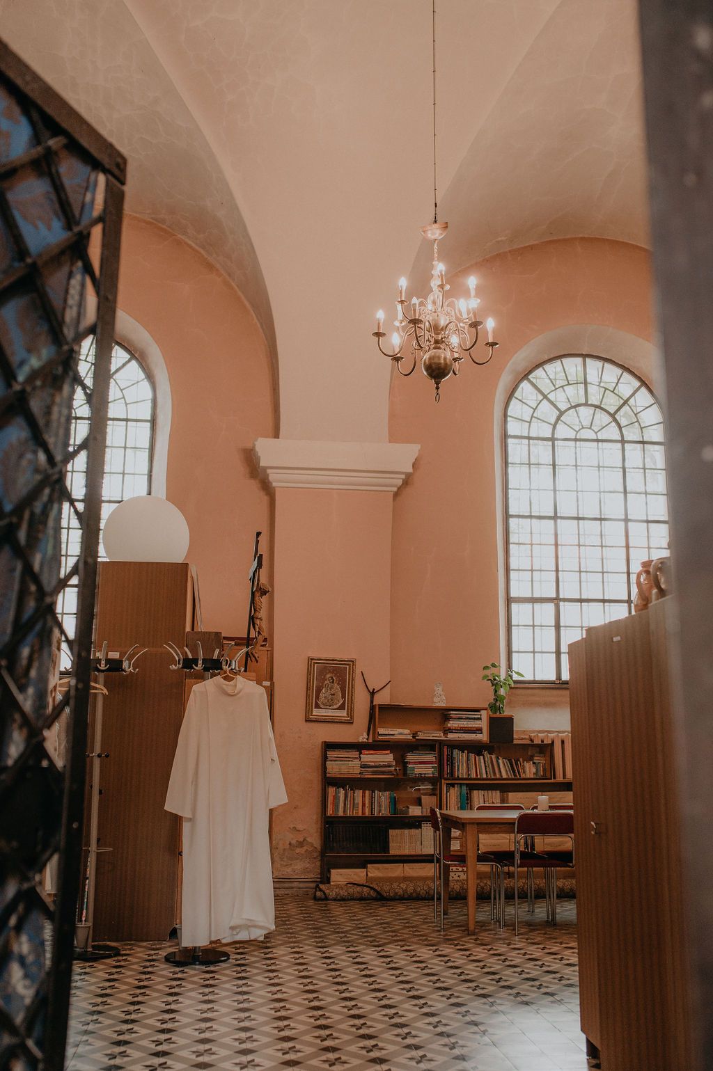 Elegant room with chandelier, arched windows, white robe on hanger, bookshelf, and patterned tile floor.