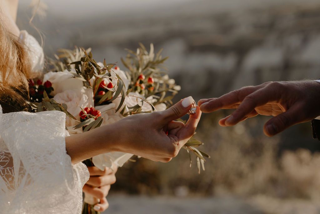 Bride and groom exchange rings surrounded by a rustic floral bouquet at golden hour.