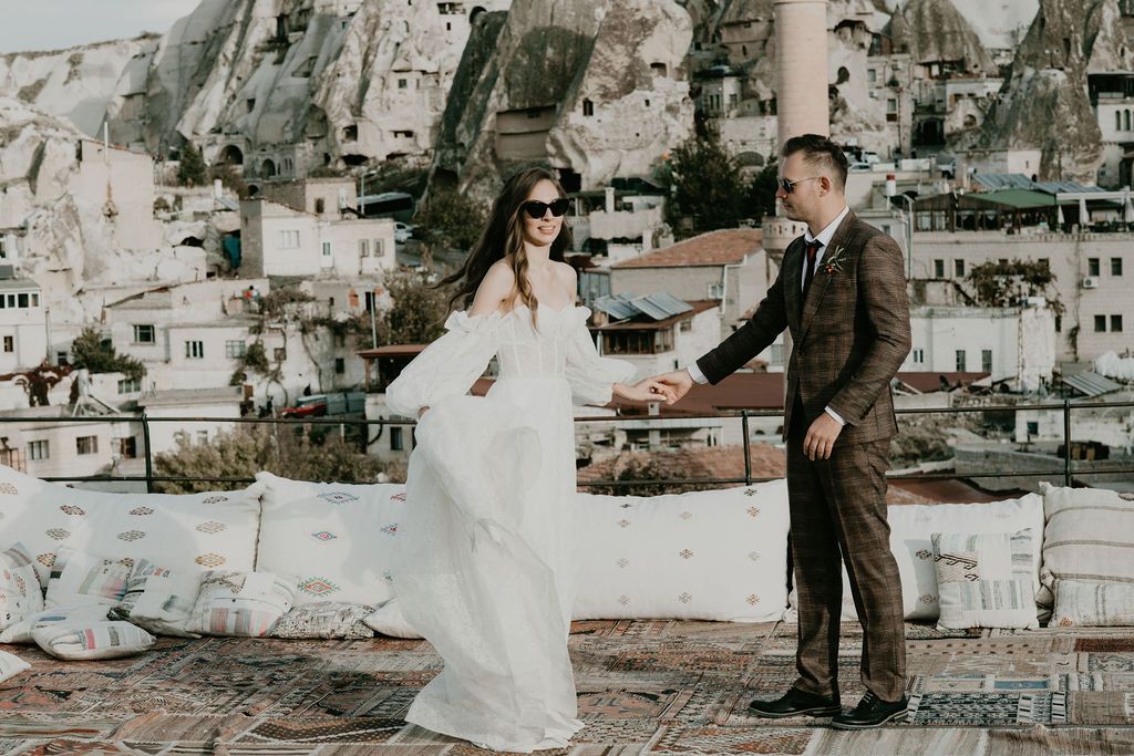 Couple in wedding attire holding hands on a rooftop in Cappadocia, Turkey, with cave dwellings in the background.