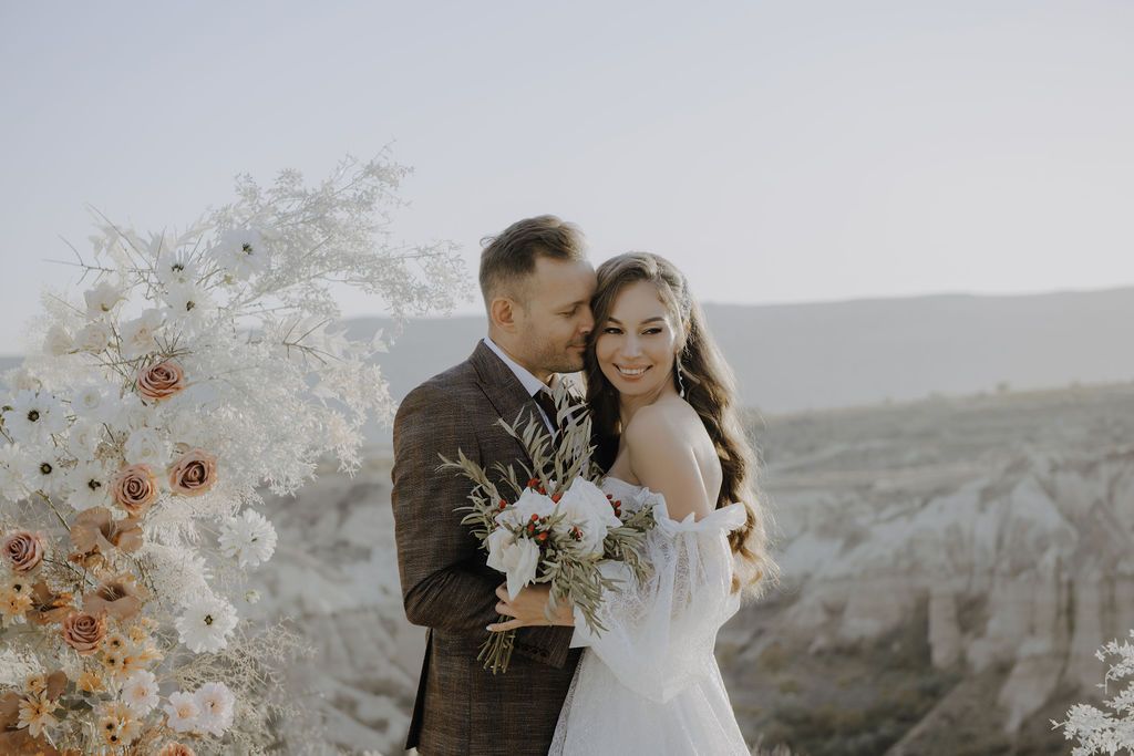 Groom tenderly kissing bride on cheek as she holds bouquet with scenic valley views at outdoor wedding.