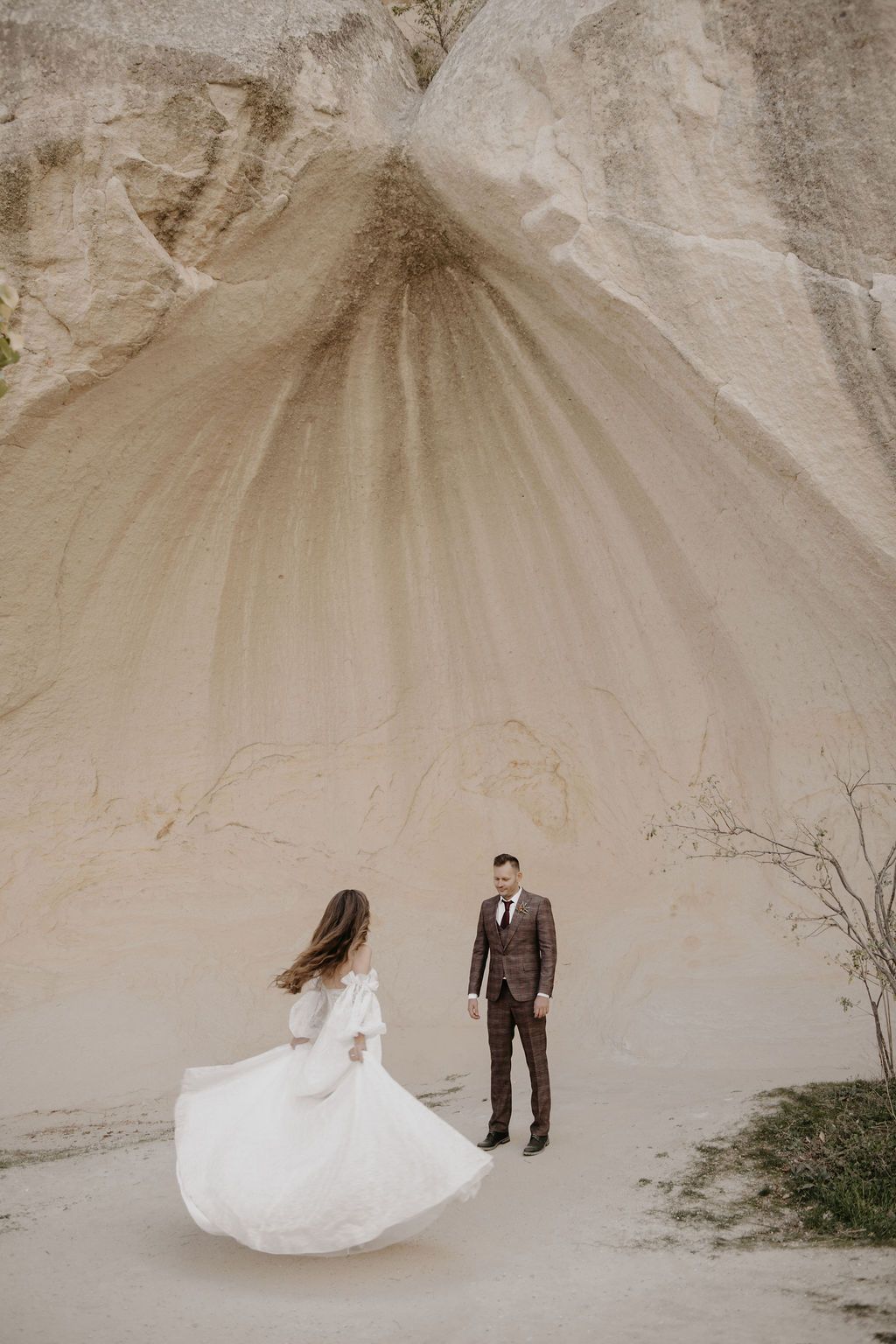 Bride in flowing white dress twirls before groom in dark suit against dramatic sandy rock formation backdrop.