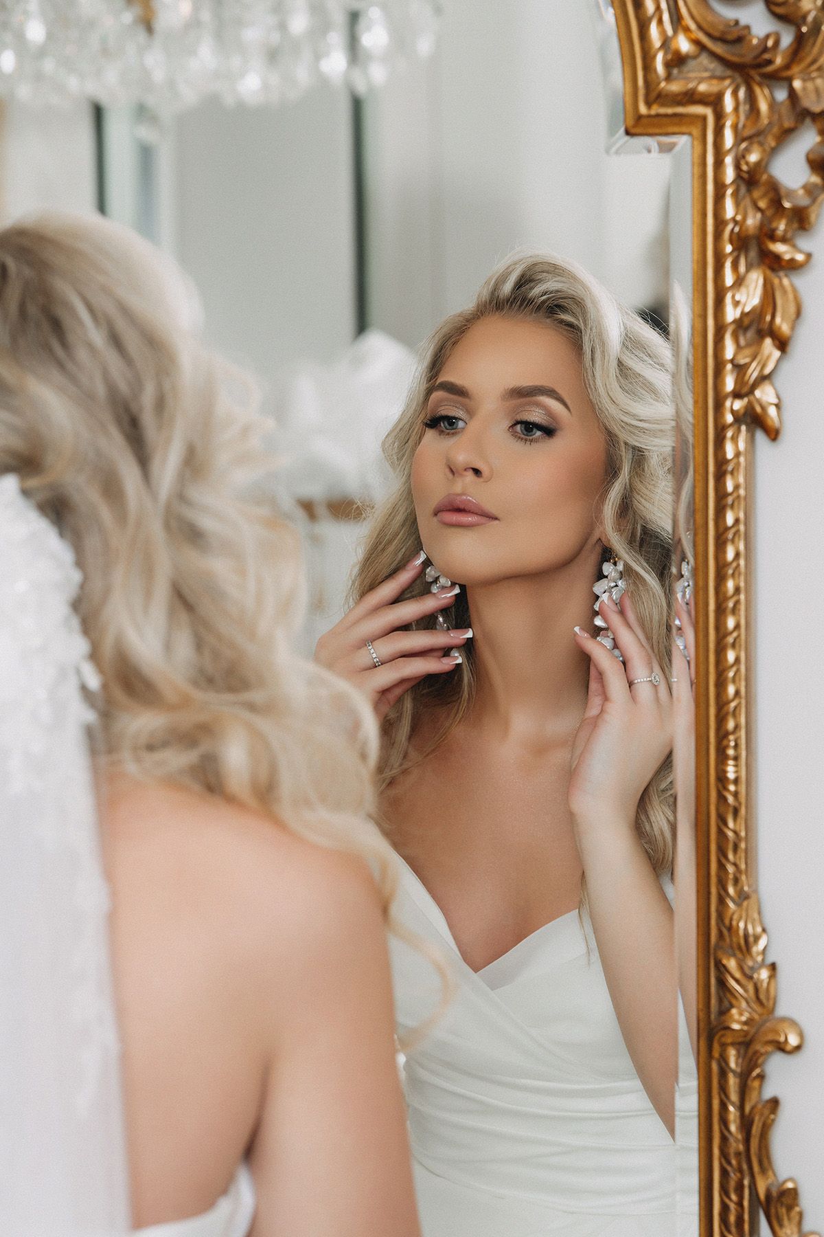 Elegant reflection in ornate gold mirror shows bride in white strapless dress with blonde hair and sparkling earrings.