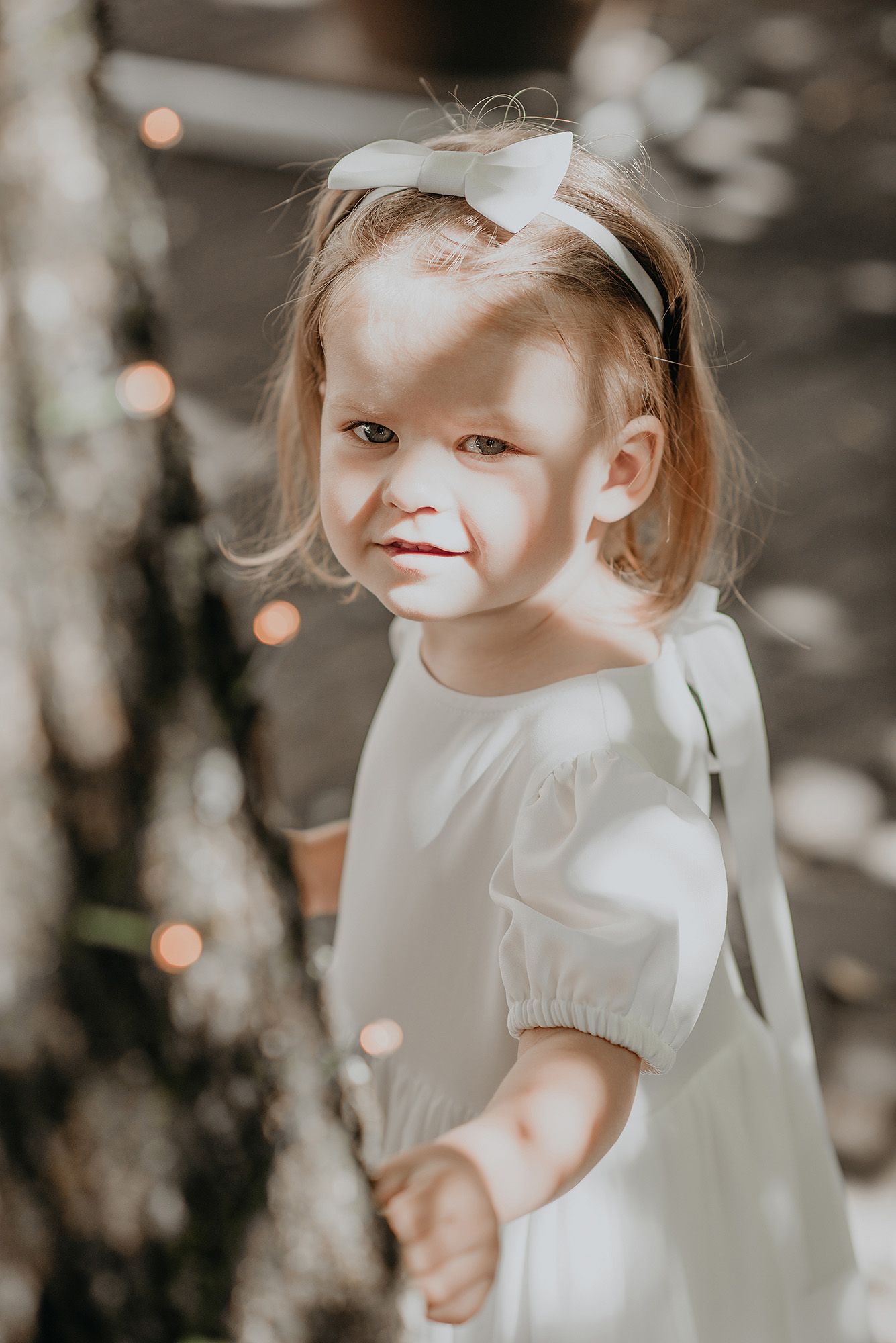 A small child in a white dress with a bow stands outdoors with bokeh lights in the background.