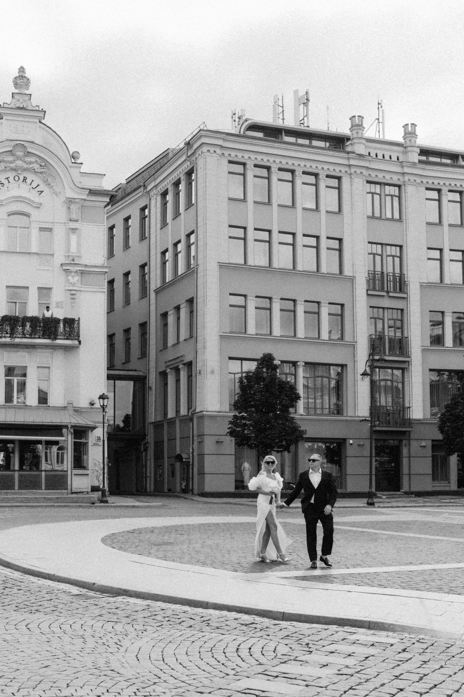 Black and white photo of a couple in elegant attire walking across a cobblestone town square in front of historic European-style buildings.