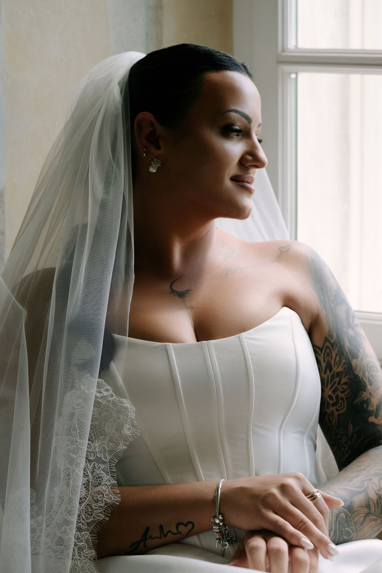 Bride in a strapless white corset gown and lace-trimmed veil gazes out a window, showing her floral arm tattoos in soft natural light.