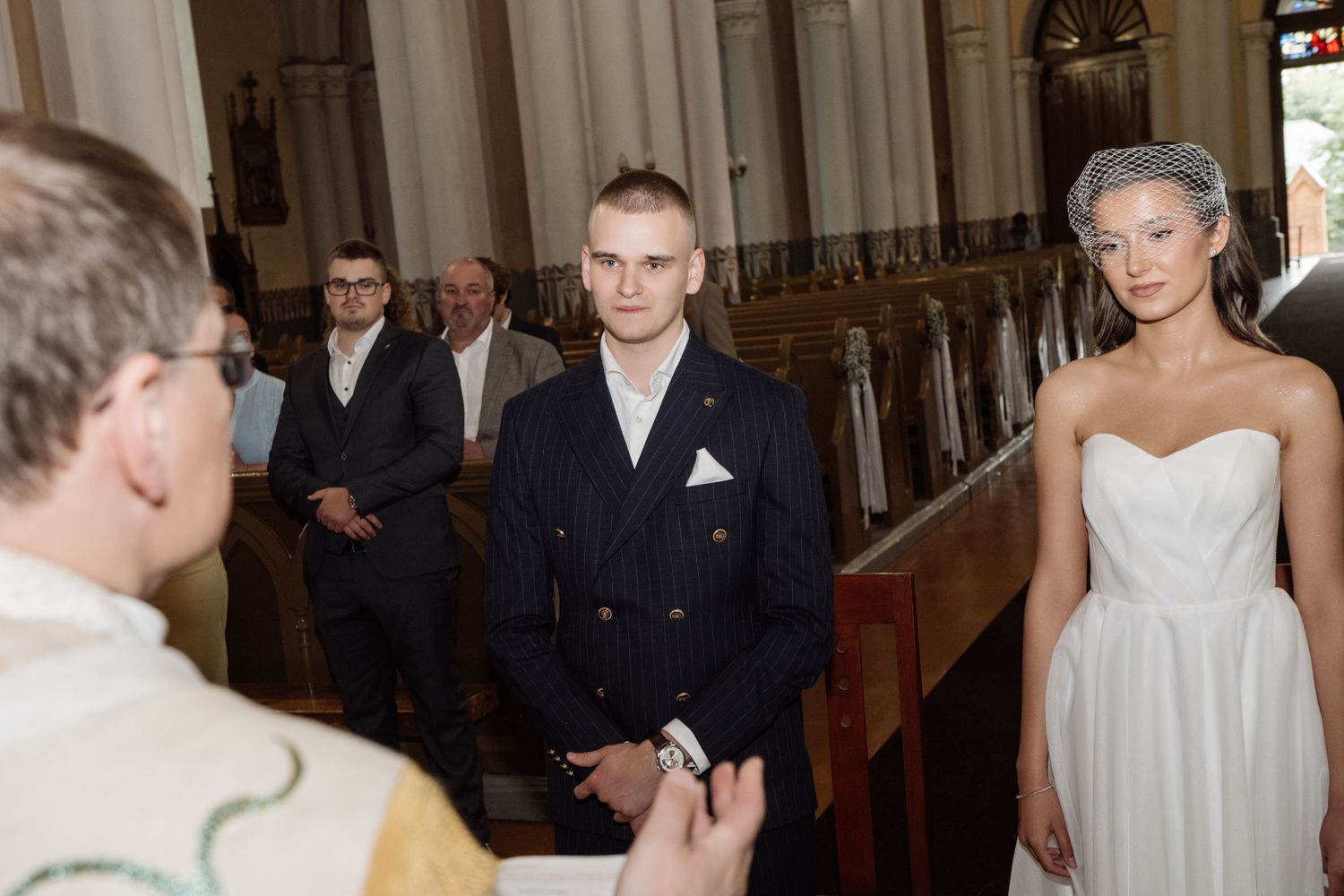 Groom in a navy pinstripe suit stands at the church altar as his bride in a white strapless gown and birdcage veil walks toward him.