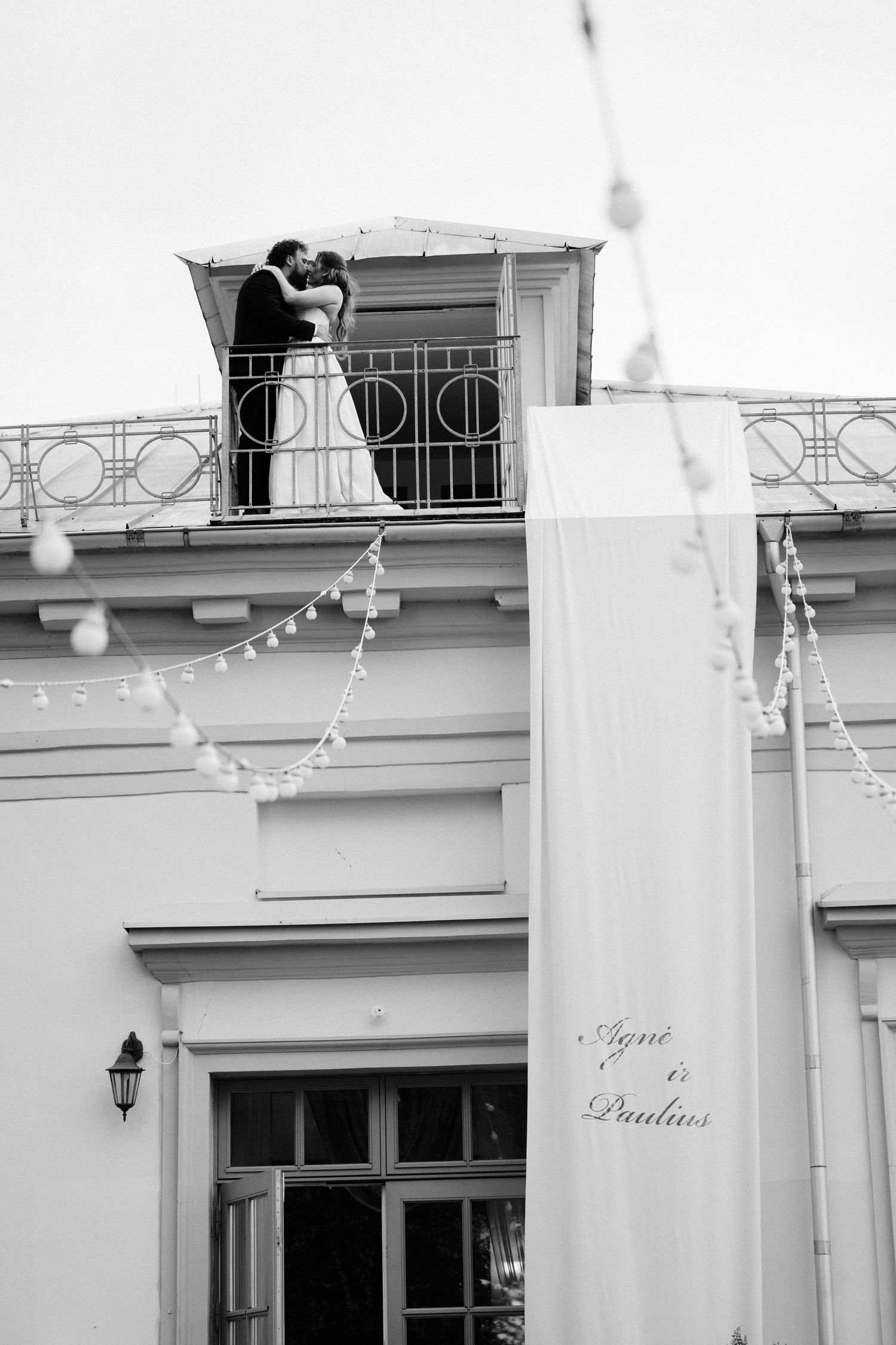 Black and white photo of newlyweds kissing on a balcony with wedding banner reading Agnes and Paulius below.