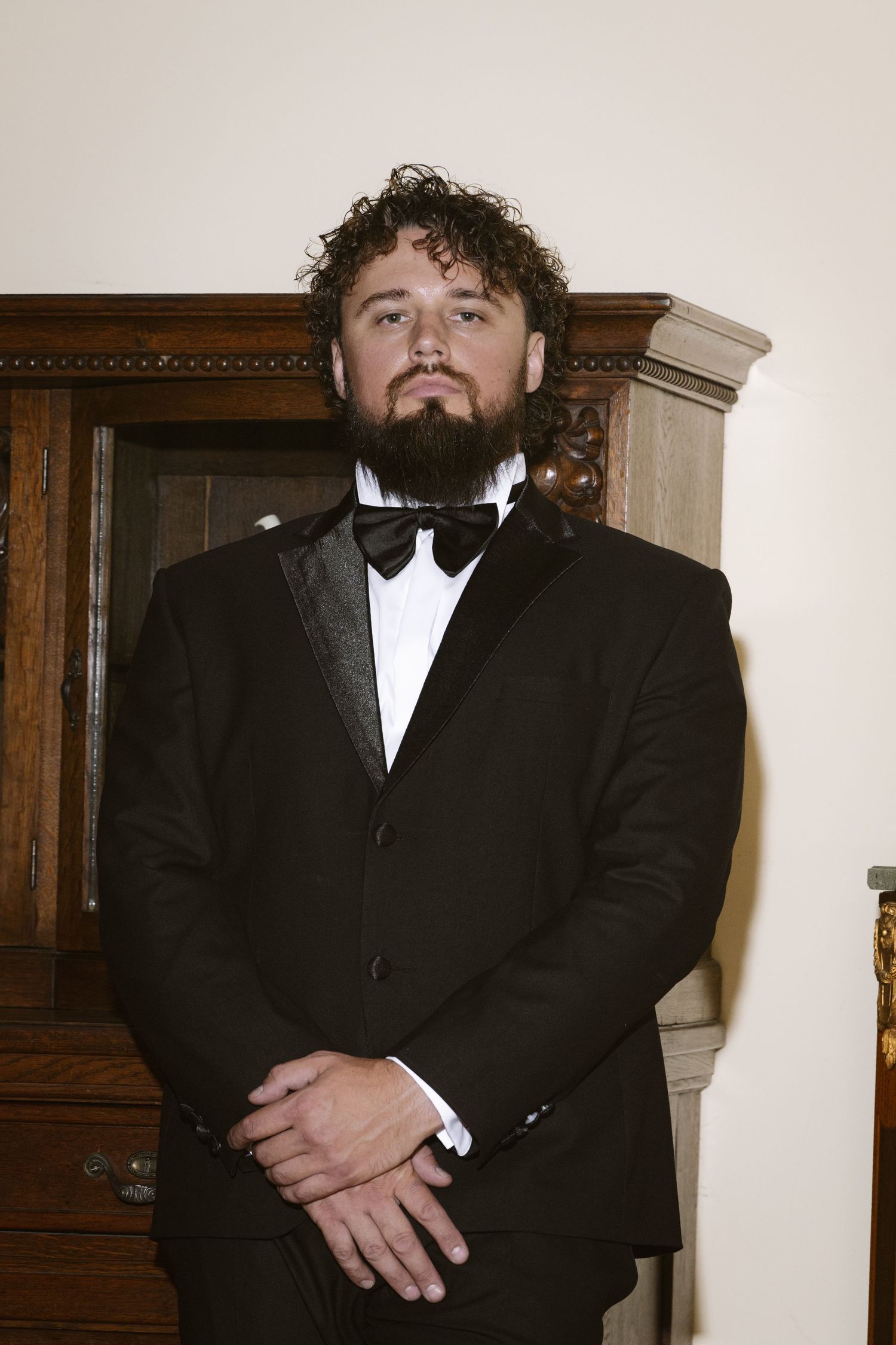 Bearded man in black tuxedo with bow tie stands confidently before wooden cabinet.