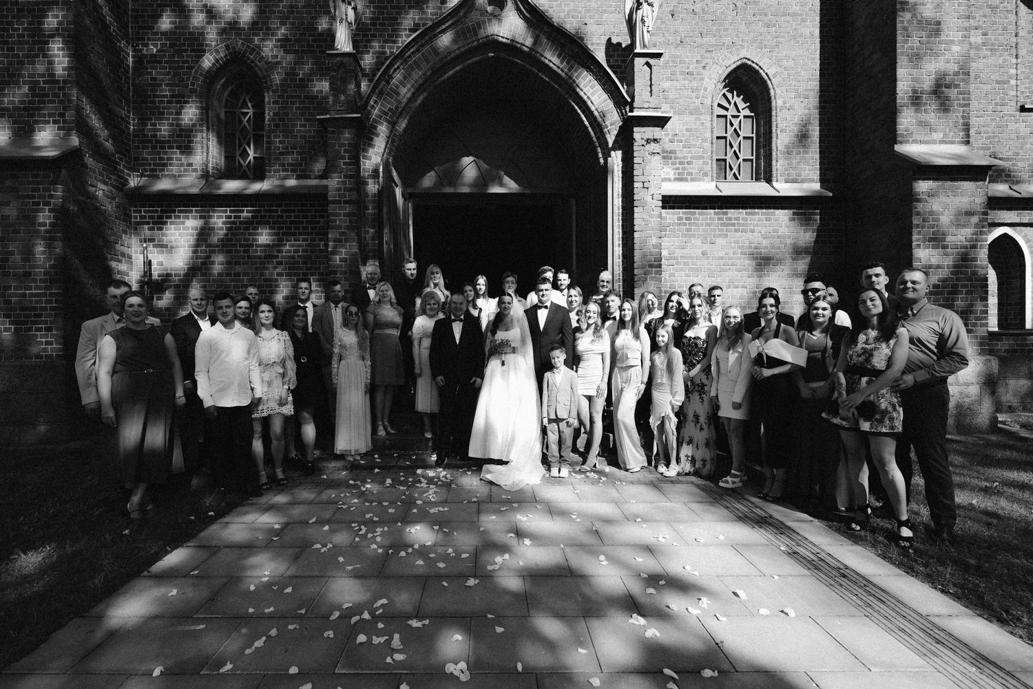 Large wedding party poses for a black and white group photo on a sunlit brick path in front of a gothic church entrance.