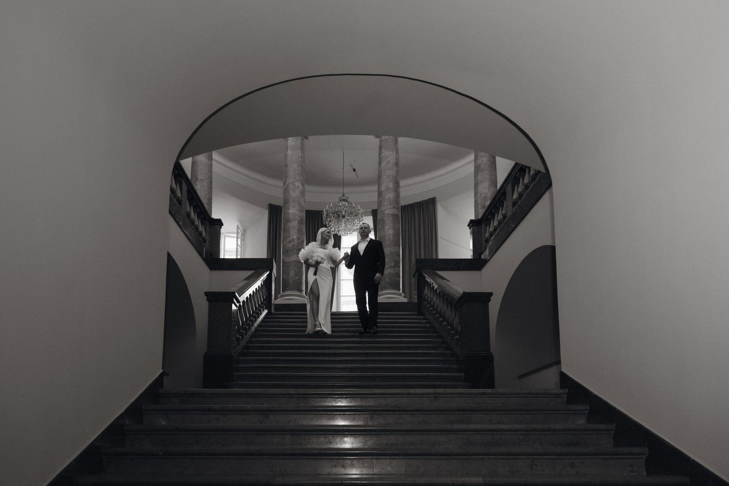 Bride in a white gown and groom in a dark suit stand together at the top of a grand marble staircase in a black and white wedding photo.