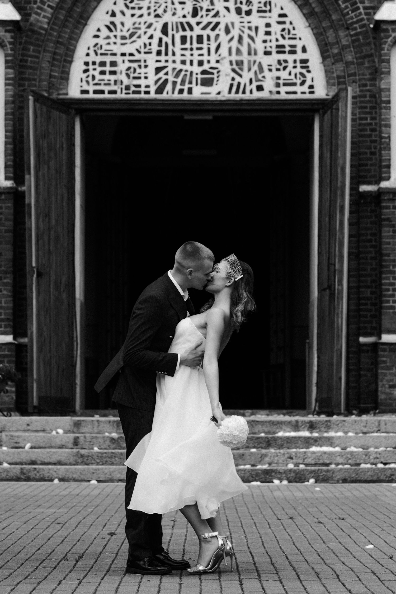 Black and white photo of a bride and groom sharing a dipped kiss on the steps of a brick church with an arched stained glass window.