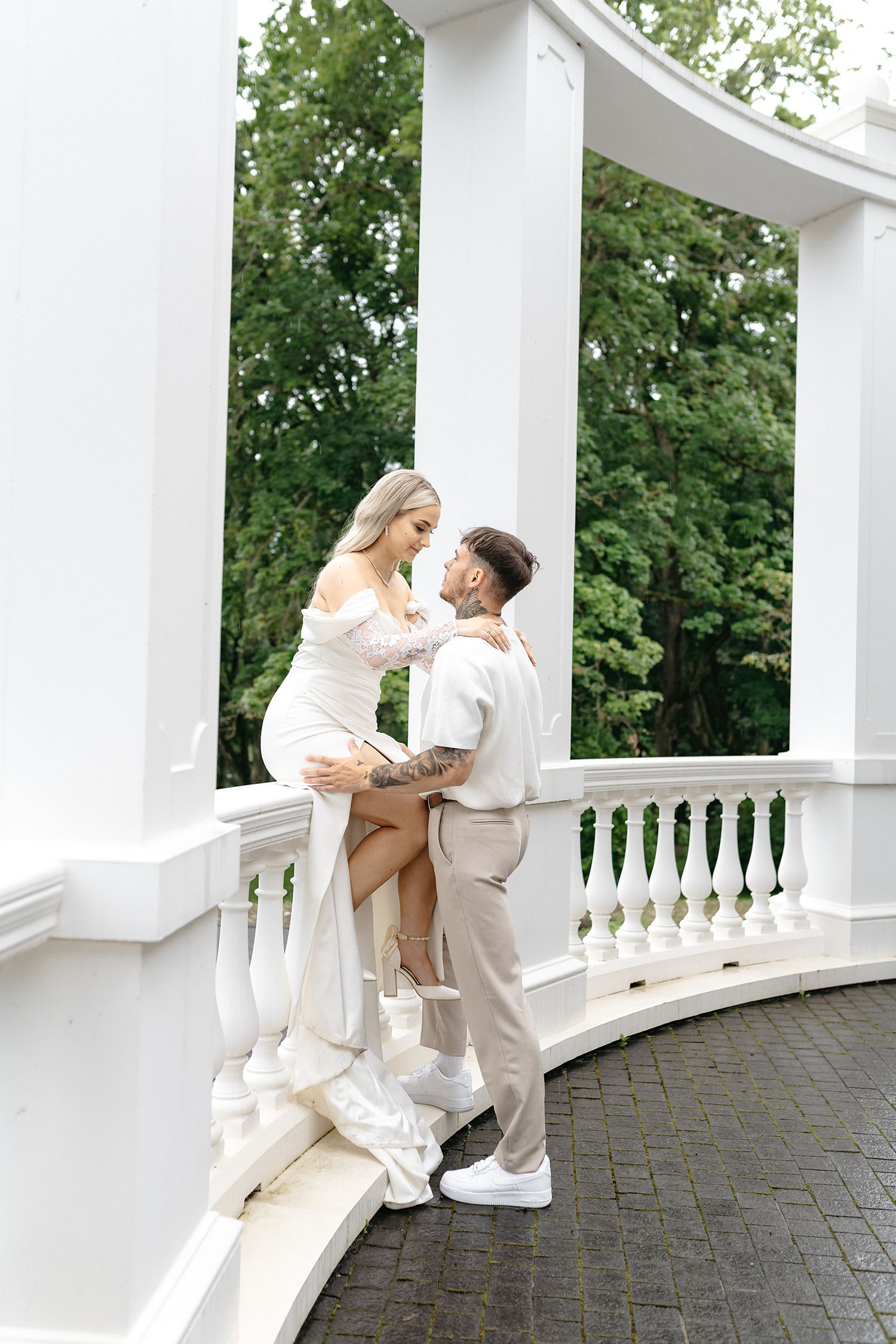 A romantic couple in white attire dancing together on an elegant columned balcony surrounded by greenery.