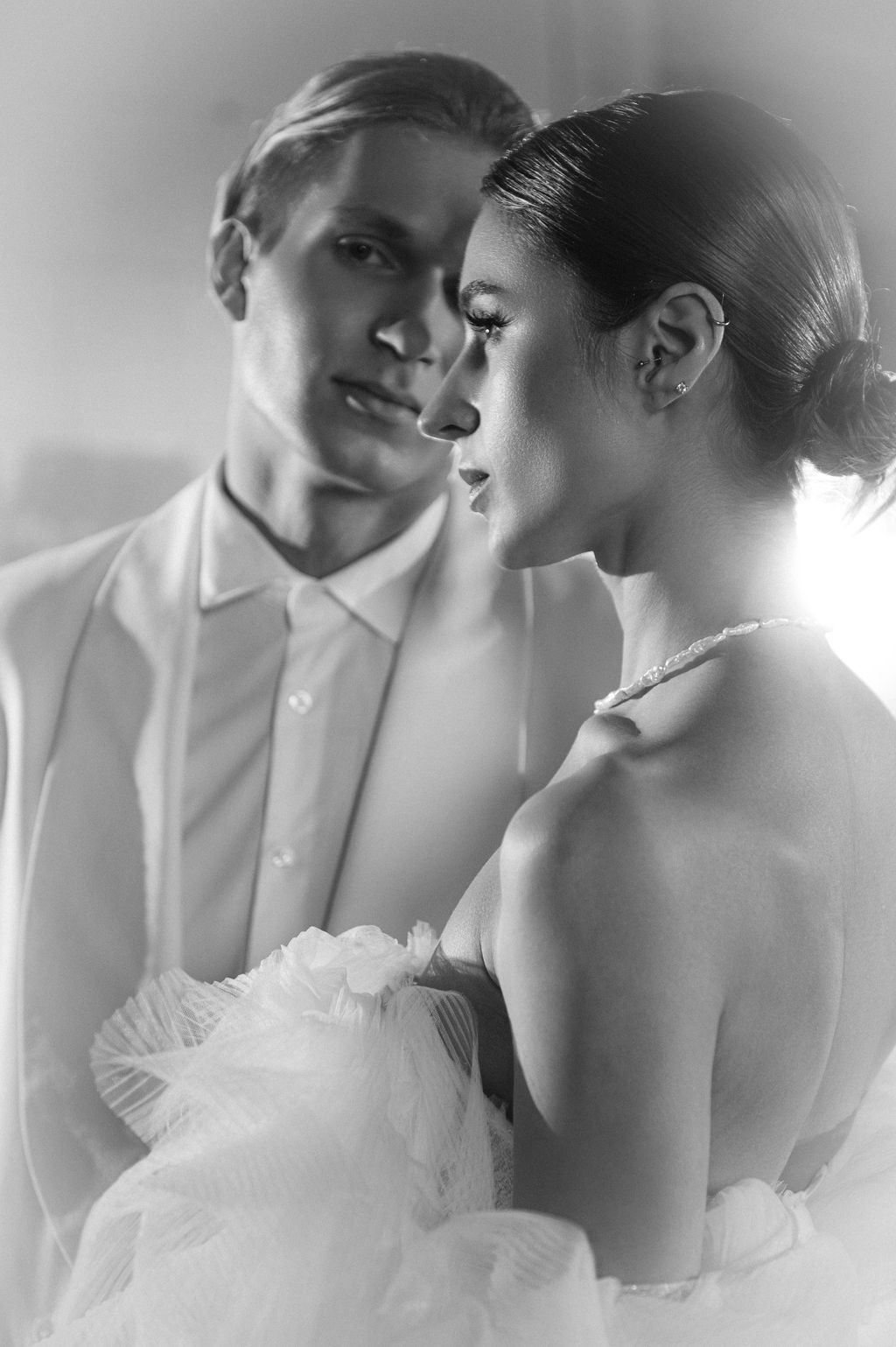 Black and white photo of an elegant couple in formal wedding attire, man in white suit gazing at woman in strapless gown.