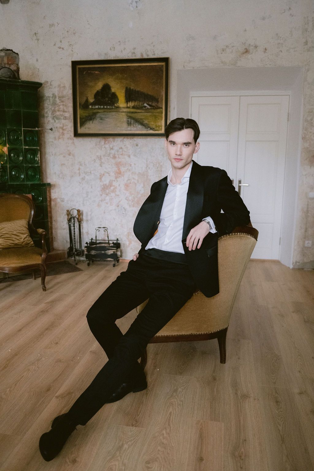 Young man in black tuxedo sitting elegantly on a vintage chair in a classic interior with painting on wall.