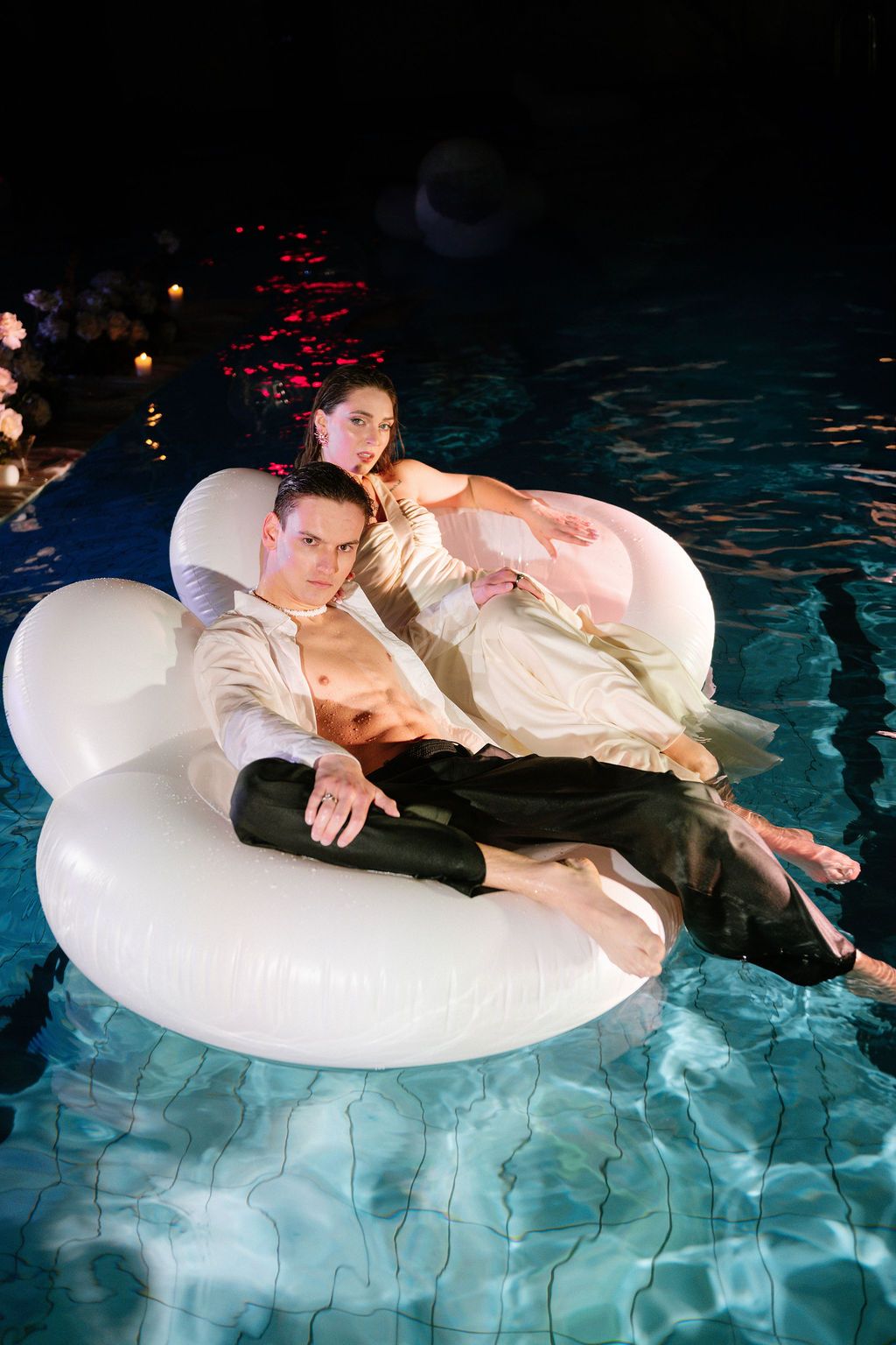 Couple relaxing on white inflatable swan float in illuminated pool at night, man shirtless in black pants, woman in elegant dress.