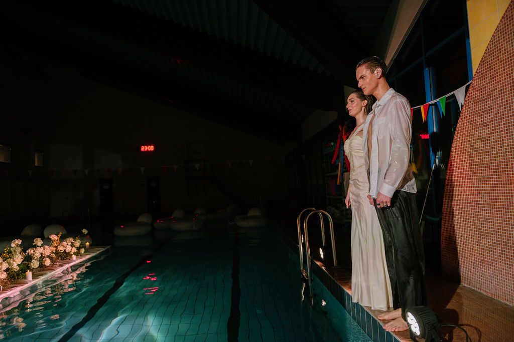 Couple standing romantically by an indoor pool at night, decorated with floating flowers and colorful string lights.
