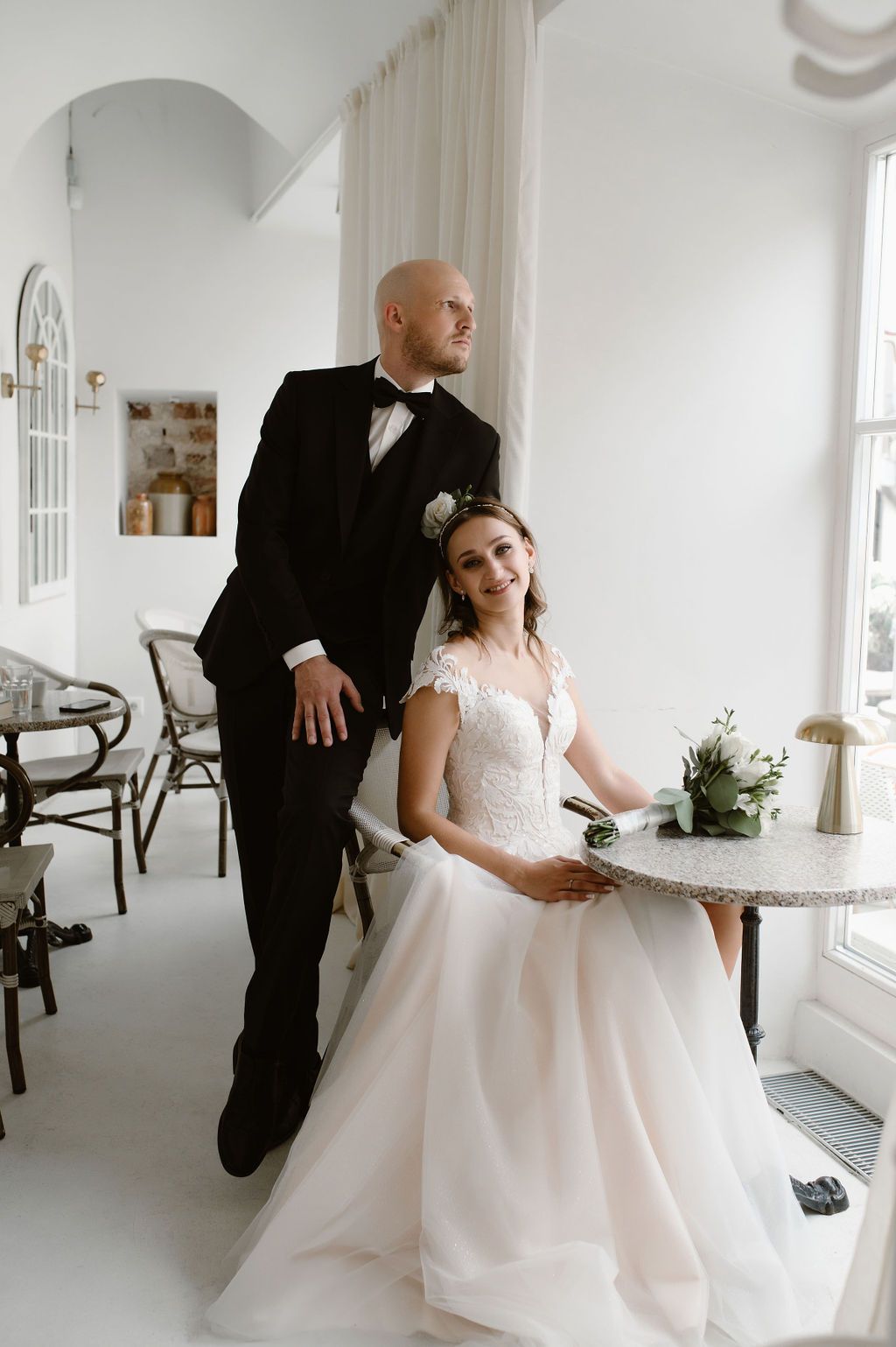 Bride in lace gown sits at table while tuxedo-clad groom stands beside her in elegant white venue.