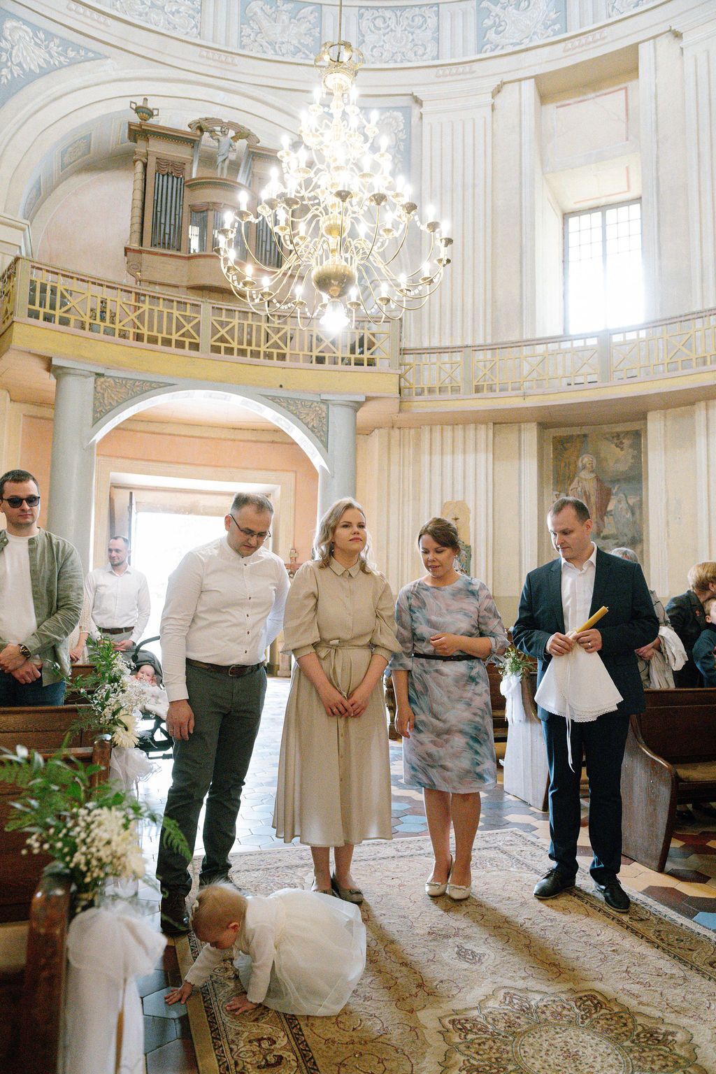 Adults and a child gather in an elegant domed church with chandelier during a baptism ceremony.