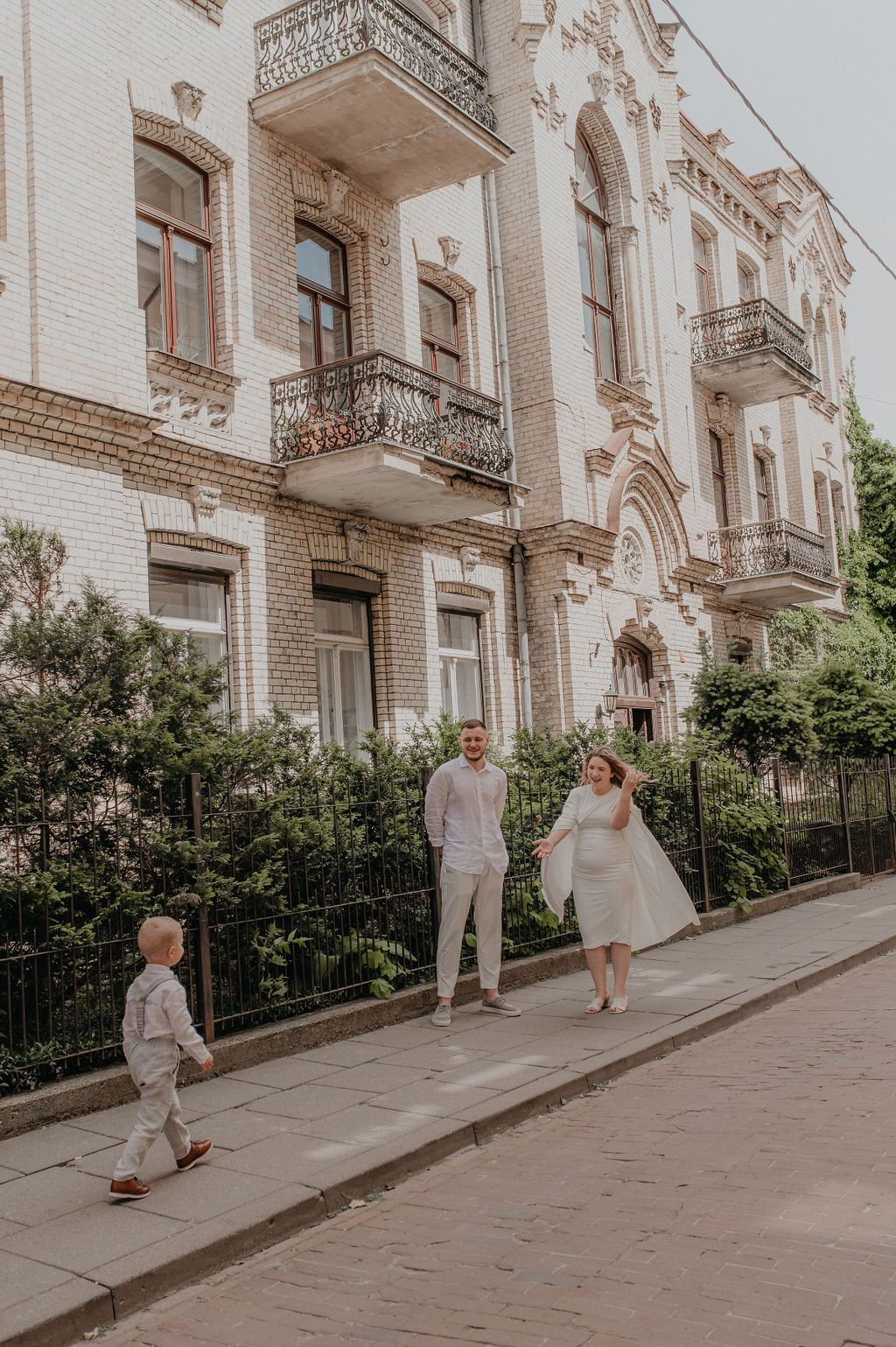 Family in white outfits walking on a sidewalk in front of an ornate historic European building with balconies.