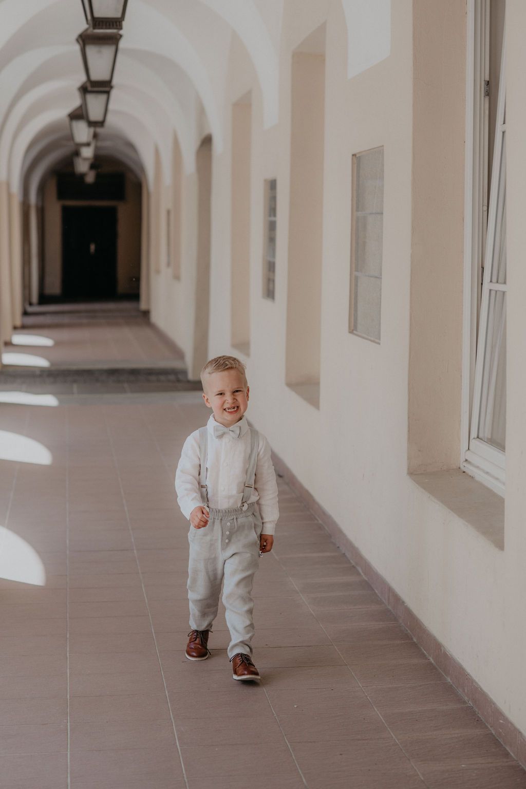 Young boy in white suspender outfit walks through sunlit arched corridor, smiling joyfully.
