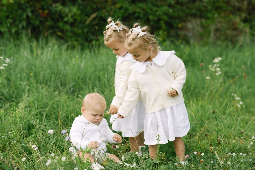 Three young girls in white dresses playing in a green meadow with wildflowers on a sunny day.