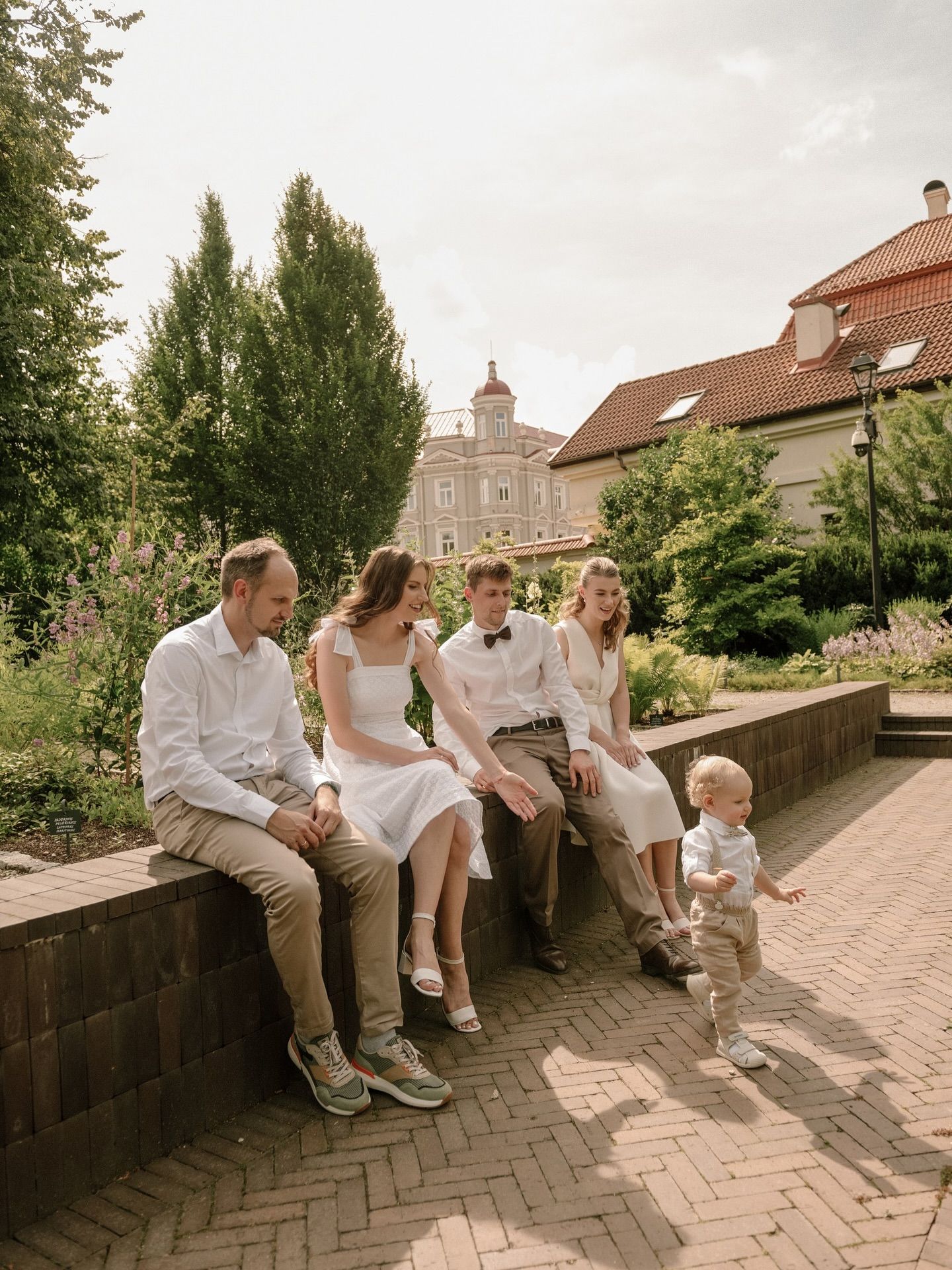 Family in white attire sitting on a brick wall in a lush garden with a historic building backdrop.