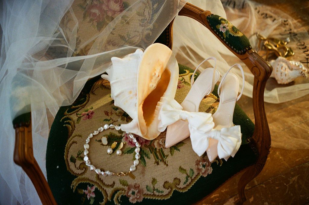 Elegant wedding accessories on ornate chair, featuring white satin heels, pearl bracelet, and bridal veil draped softly.