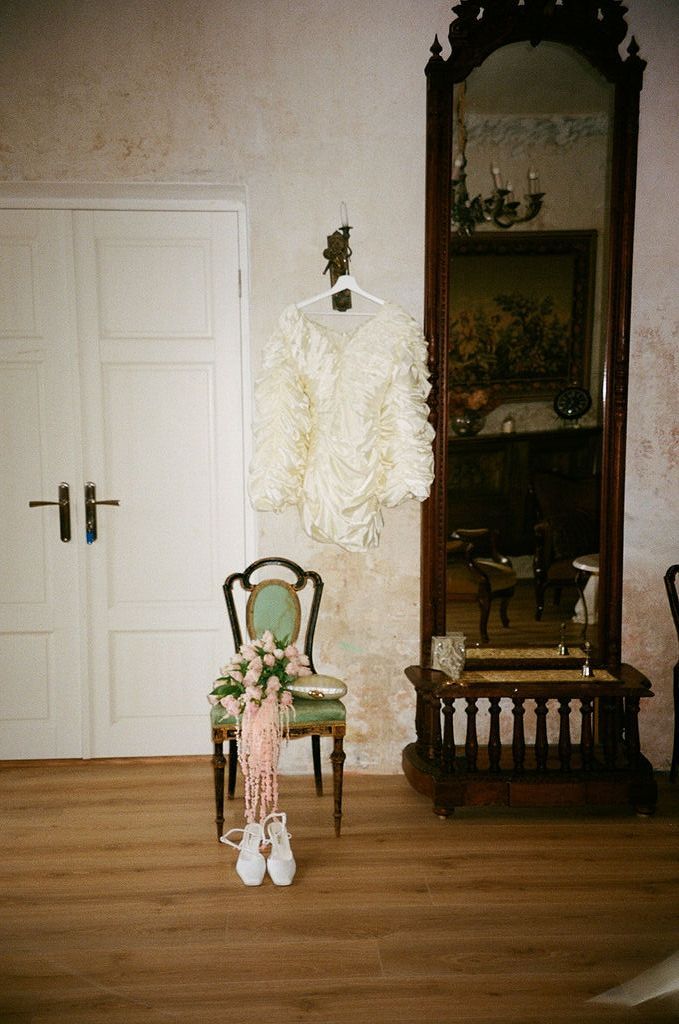 Elegant wedding dress hanging on antique green chair beside ornate floor mirror in vintage room with hardwood floors.