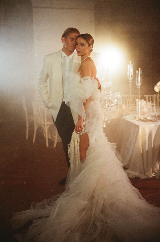 Couple in wedding attire pose romantically in dramatic backlit reception hall, bride in mermaid gown with tulle.